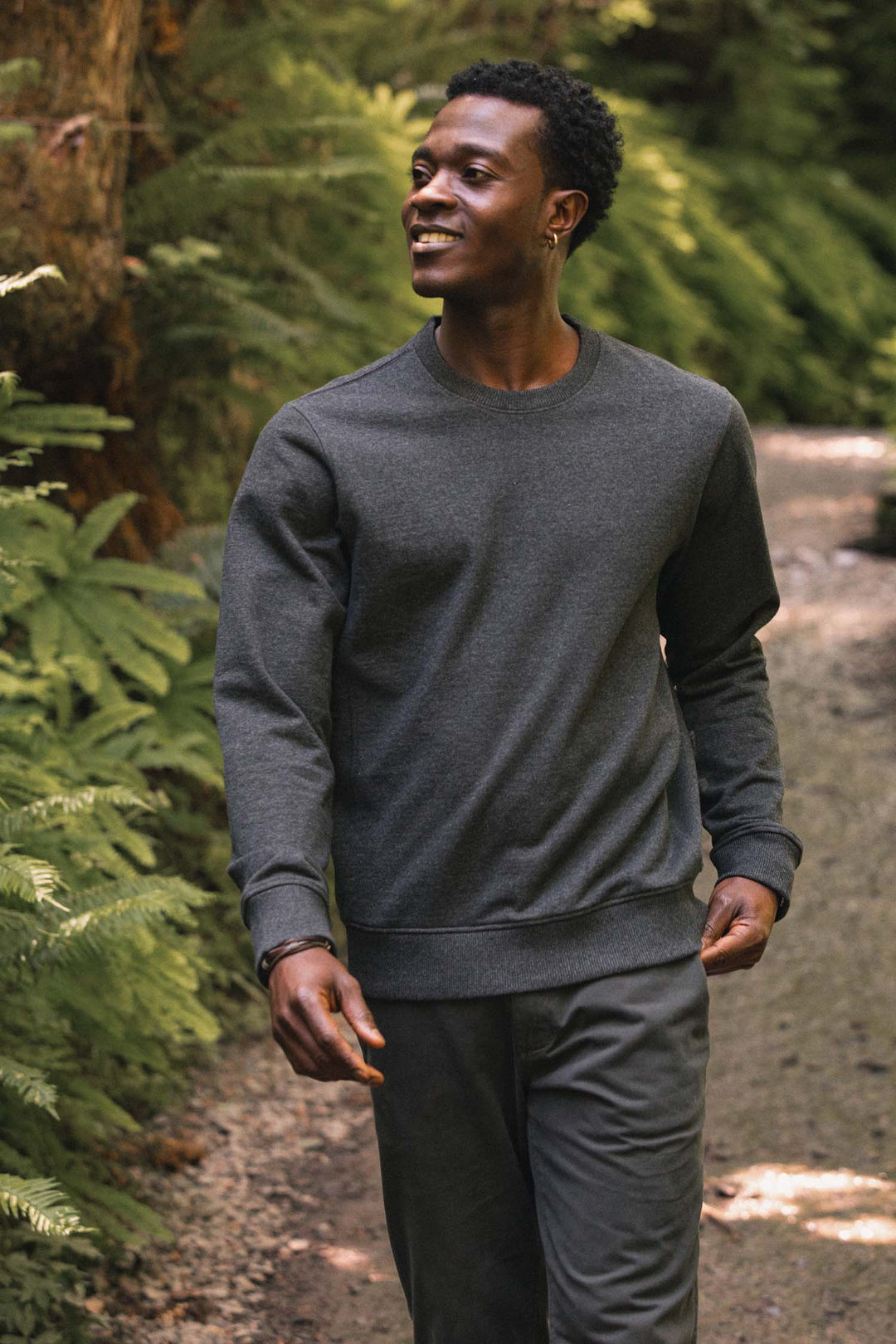 A man wearing the Bridge & Burn Hayden Crew Sweatshirt in Graphite with matching pants walks along a forest path, smiling amid green foliage and sunlight.