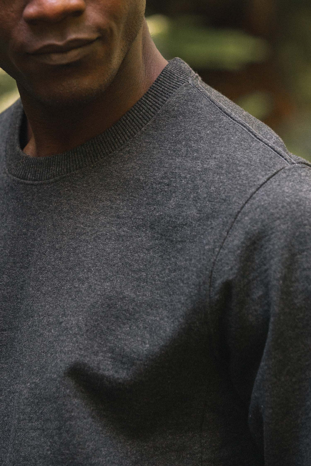 A close-up of a person wearing the Bridge & Burn Hayden Crew Sweatshirt in Graphite, crafted from recycled cotton, with only the lower half of their face visible and ribbed cuffs accenting the sleeves against a blurred outdoor background.