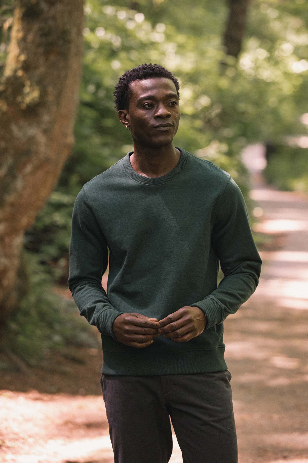 A man wearing the Bridge & Burn Hayden Crew Sweatshirt in Juniper stands on a forest path, holding something in his hands as sunlight filters through the trees behind him.