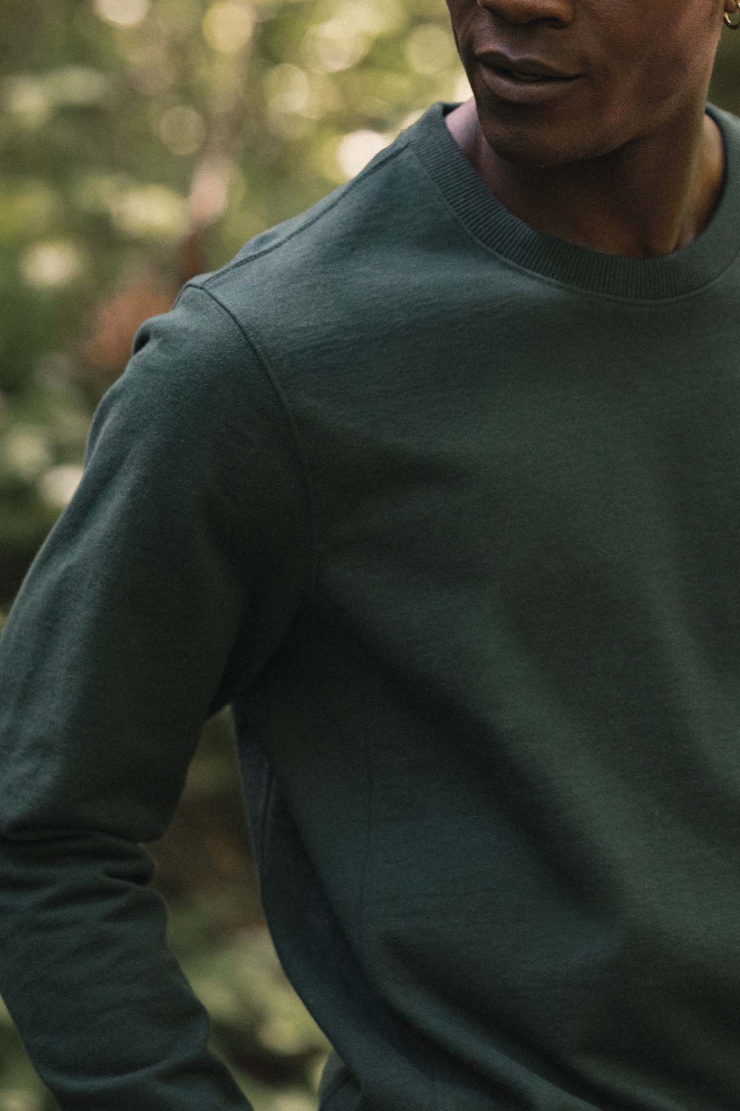 A person wearing the Bridge & Burn Hayden Crew Sweatshirt in Juniper, a dark green long-sleeve crew neck made from recycled cotton, stands outdoors with only their torso and lower face visible against a backdrop of blurred greenery.