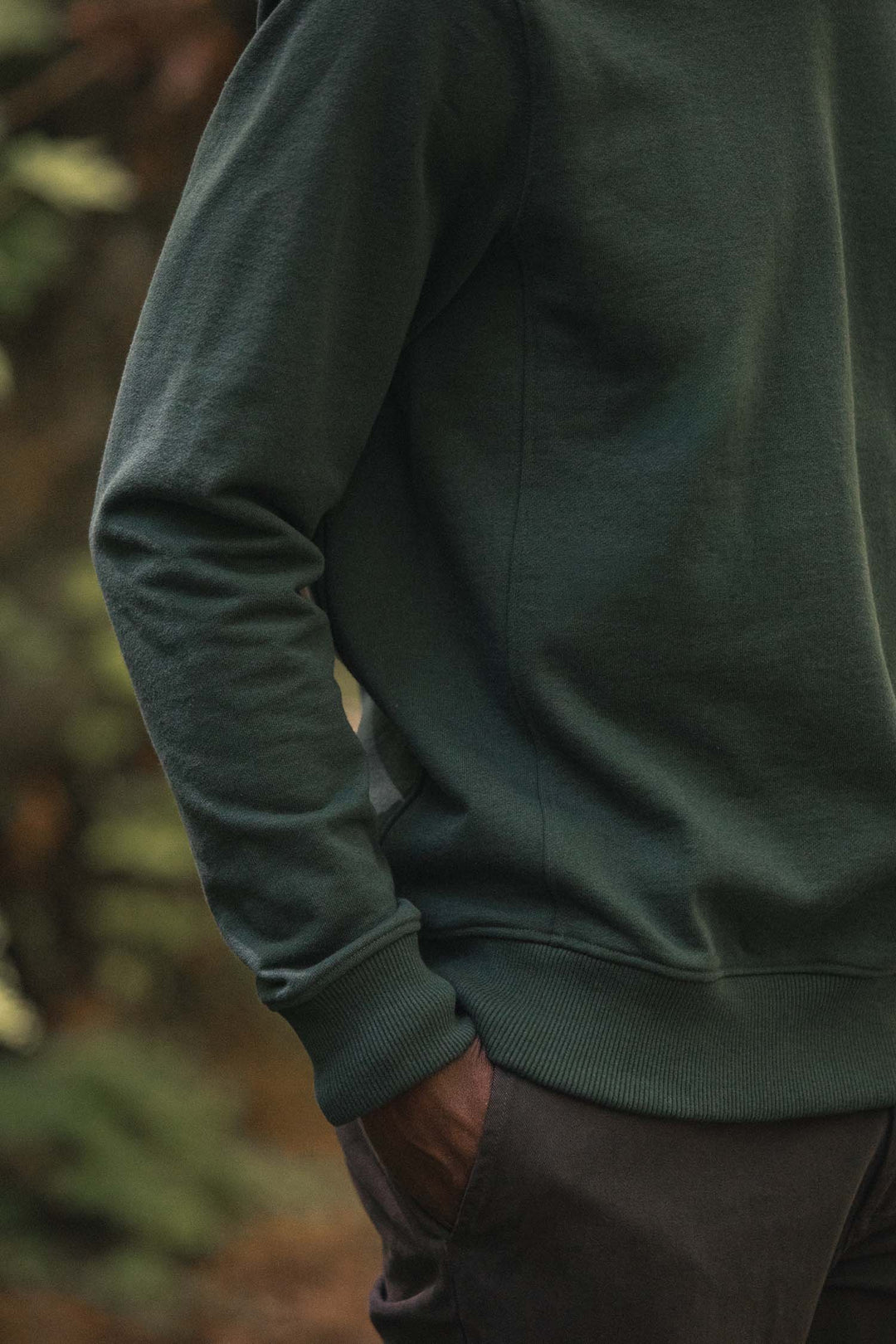 A person wearing the Bridge & Burn Hayden Crew Sweatshirt in Juniper and dark pants stands outdoors with one hand in their pocket, surrounded by blurred greenery in the background.