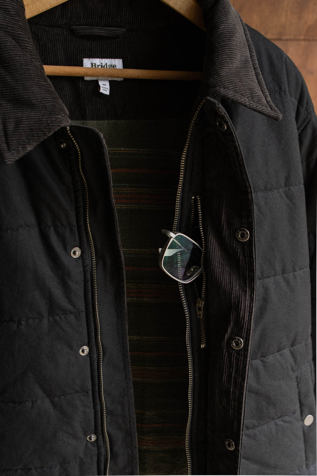 Black Bridge & Burn Holman Quilted Jacket / Black Canvas with a corduroy collar, hanging on a wooden hanger with a pair of glasses tucked into the chest zipper pocket.