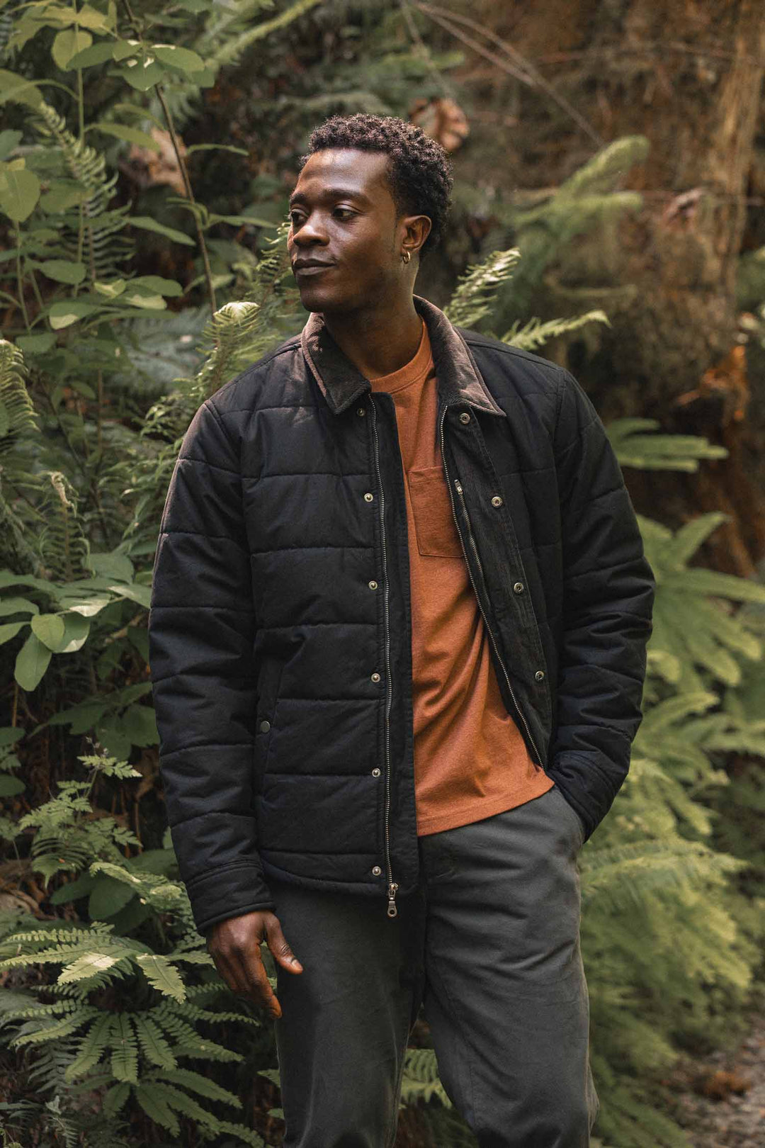 A man wearing a black Holman Quilted Jacket / Black Canvas over a burnt-orange t-shirt stands in a forest.