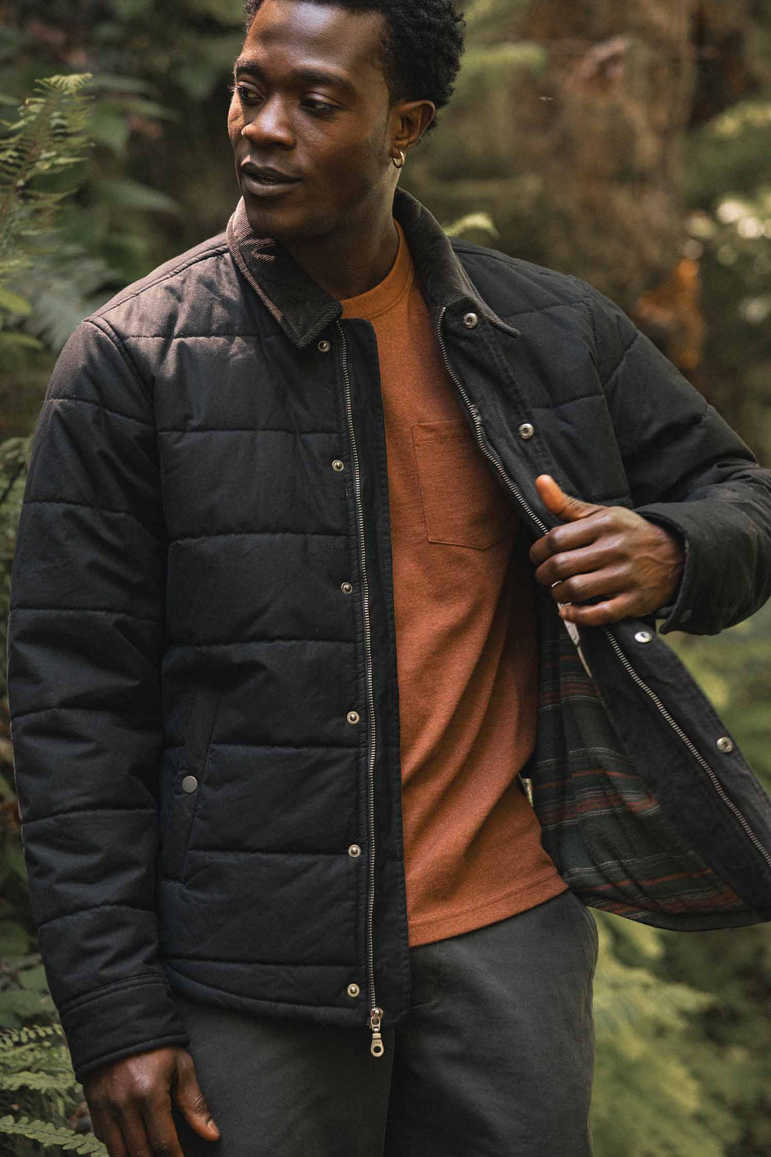 A man wearing a black Holman Quilted Jacket / Black Canvas over a burnt-orange t-shirt.