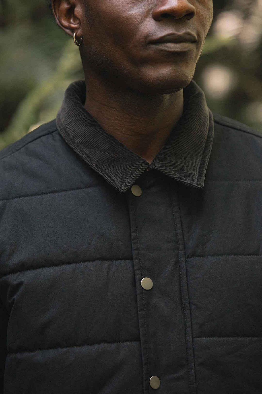 Close-up of a person wearing a black canvas Holman Quilted Jacket with a rugged quilted texture, corduroy collar, and metal snap buttons.