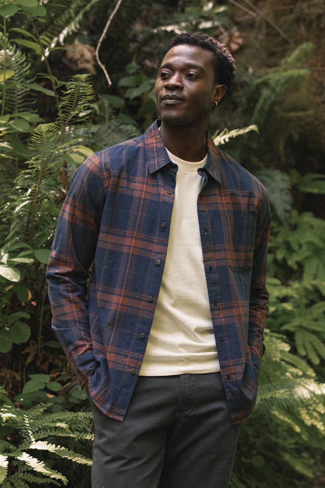 A person stands outdoors among green plants, wearing the Bridge & Burn Larson Shirt in Cinder Plaid—a blue and orange brushed flannel plaid classic fit button-up—layered over a white t-shirt and dark pants, looking slightly to the side with a calm expression.