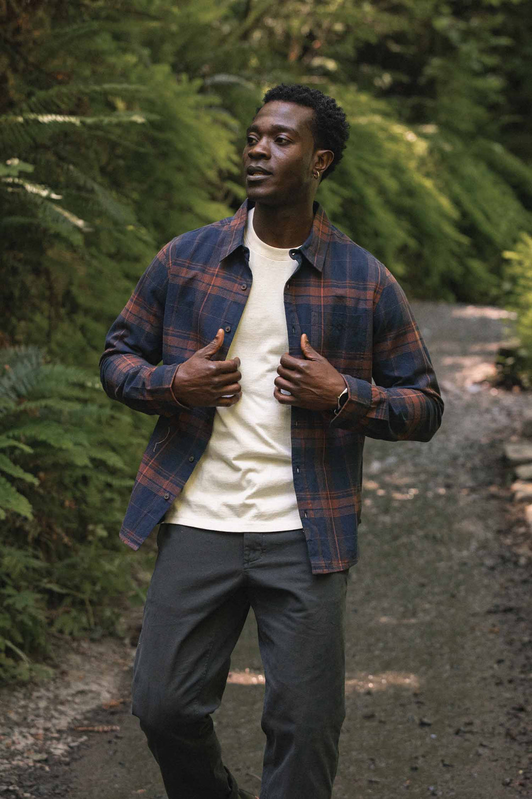 A man wearing the Bridge & Burn Larson Shirt in Cinder Plaid, layered over a white t-shirt, walks along a gravel path bordered by greenery and ferns, gazing thoughtfully to the side.