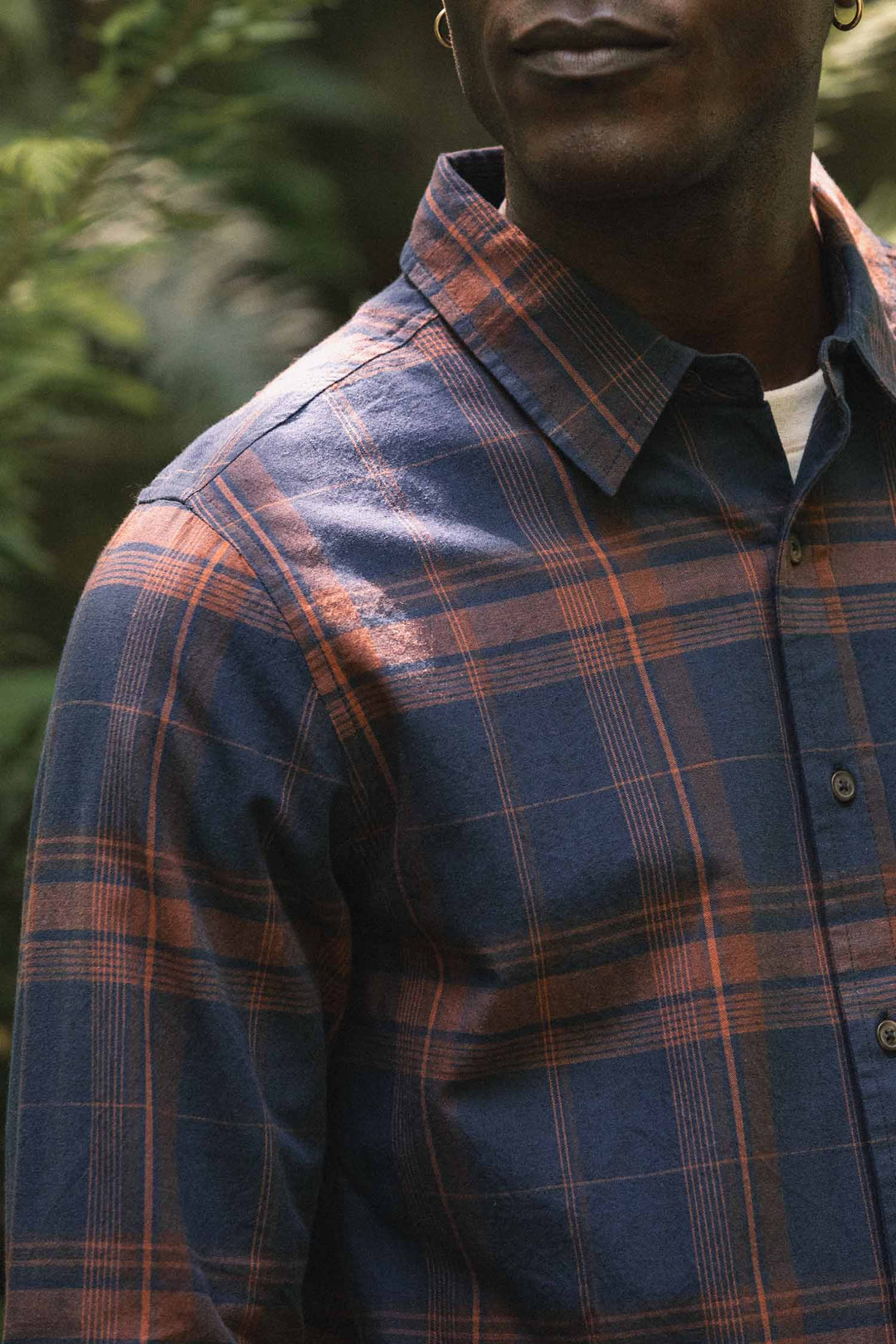 A person wearing the Bridge & Burn Larson Shirt in Cinder Plaid stands outdoors, surrounded by greenery. Only the lower half of their face and upper torso are visible.
