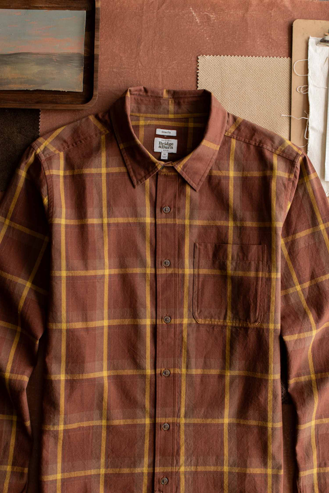 The Bridge & Burn Larson Shirt in Harvest Plaid, a classic fit brown brushed flannel button-up with a chest pocket, is laid flat on a brown surface next to fabric swatches and a small wooden frame.