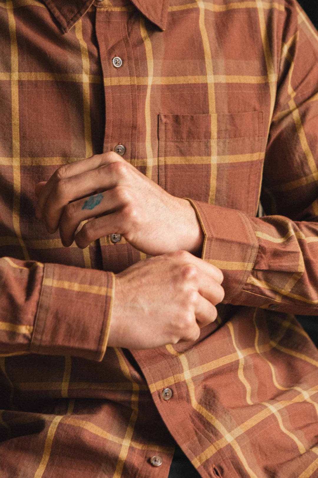 A person wearing the Bridge & Burn Larson Shirt in Harvest Plaid—a classic fit button-up brown and yellow brushed flannel—is buttoning the cuff of their sleeve. Only the torso and hands are visible.