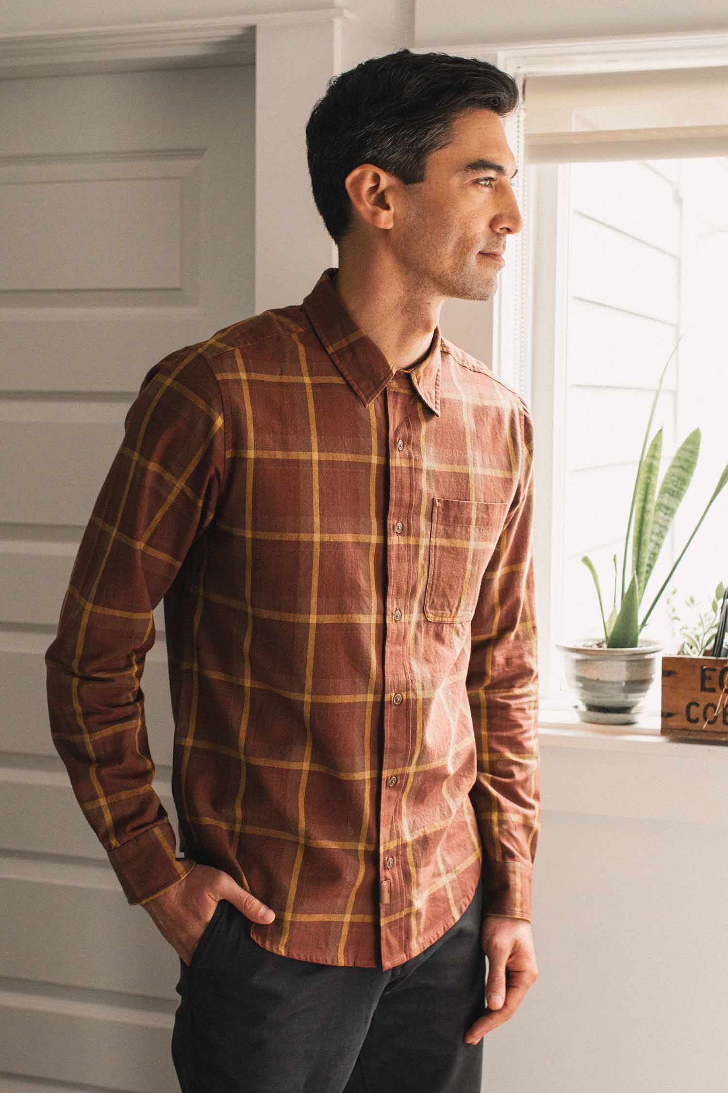 A man wearing the Bridge & Burn Larson Shirt in Harvest Plaid, a classic fit button-up brown brushed flannel, stands indoors near a window with plants on the sill, looking outside with his hand in his pocket.
