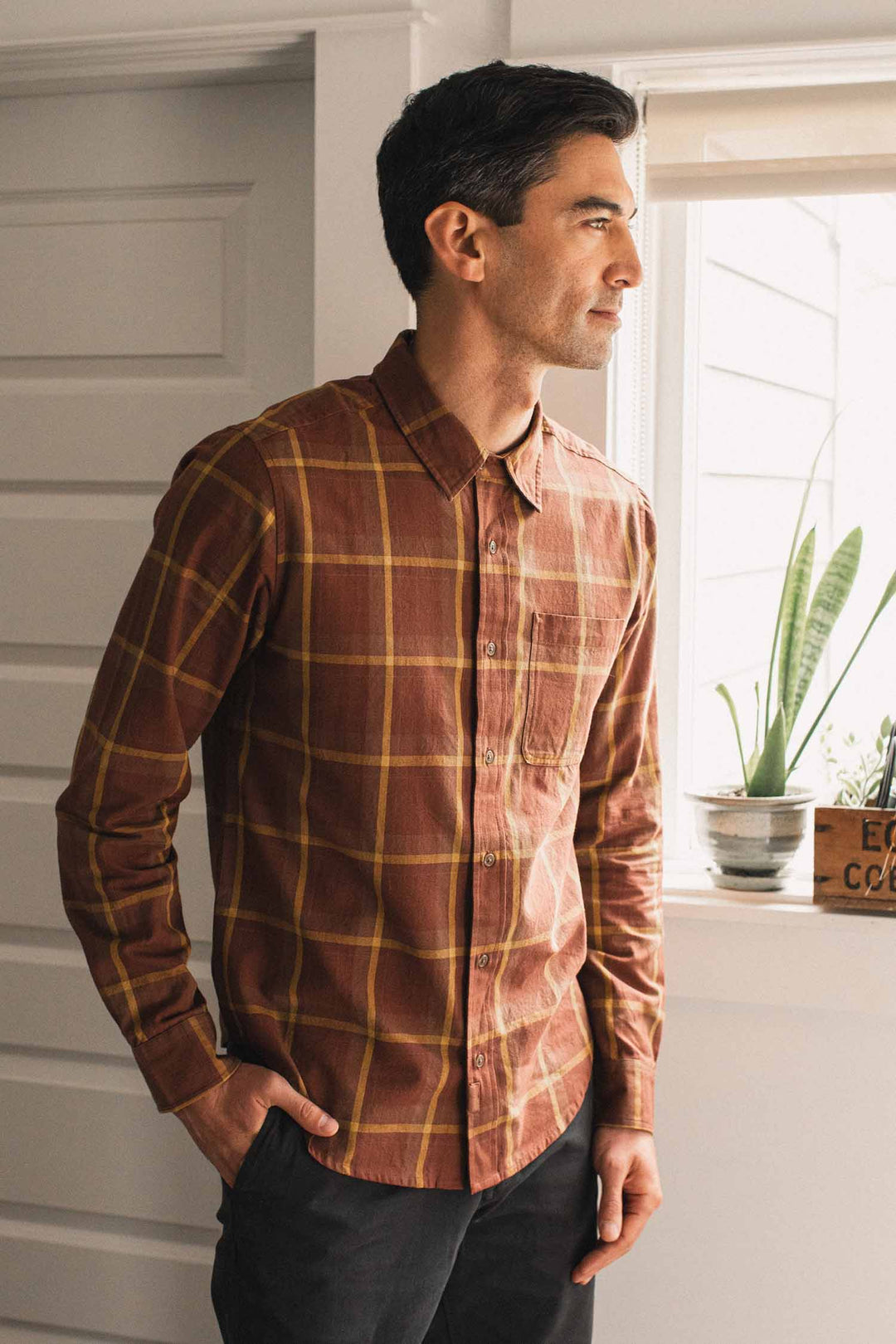 A man wearing the Bridge & Burn Larson Shirt in Harvest Plaid, a classic fit button-up brown brushed flannel, stands indoors near a window with plants on the sill, looking outside with his hand in his pocket.