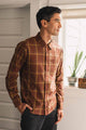A man wearing the Bridge & Burn Larson Shirt in Harvest Plaid, a classic fit button-up brown brushed flannel, stands indoors near a window with plants on the sill, looking outside with his hand in his pocket.