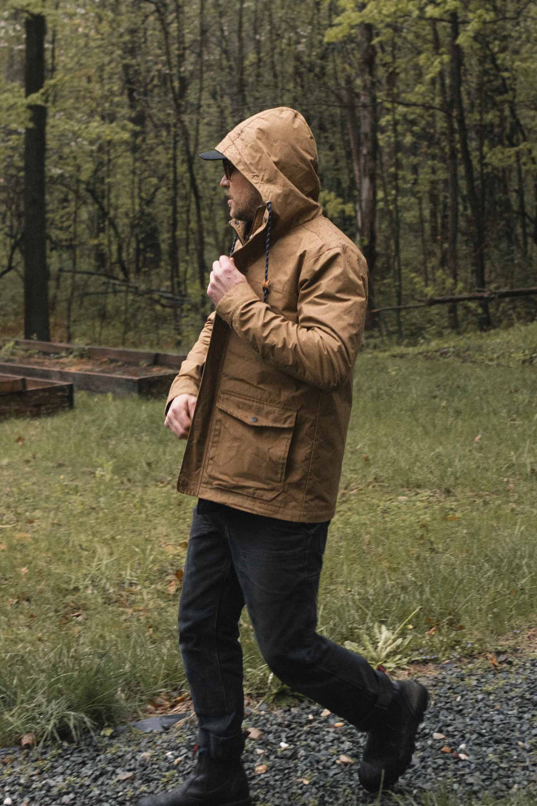 A person wearing a hooded, water-resistant Bridge & Burn Marshall Jacket / Dark Khaki Waxed Canvas and dark jeans walks along a gravel path in a forest.