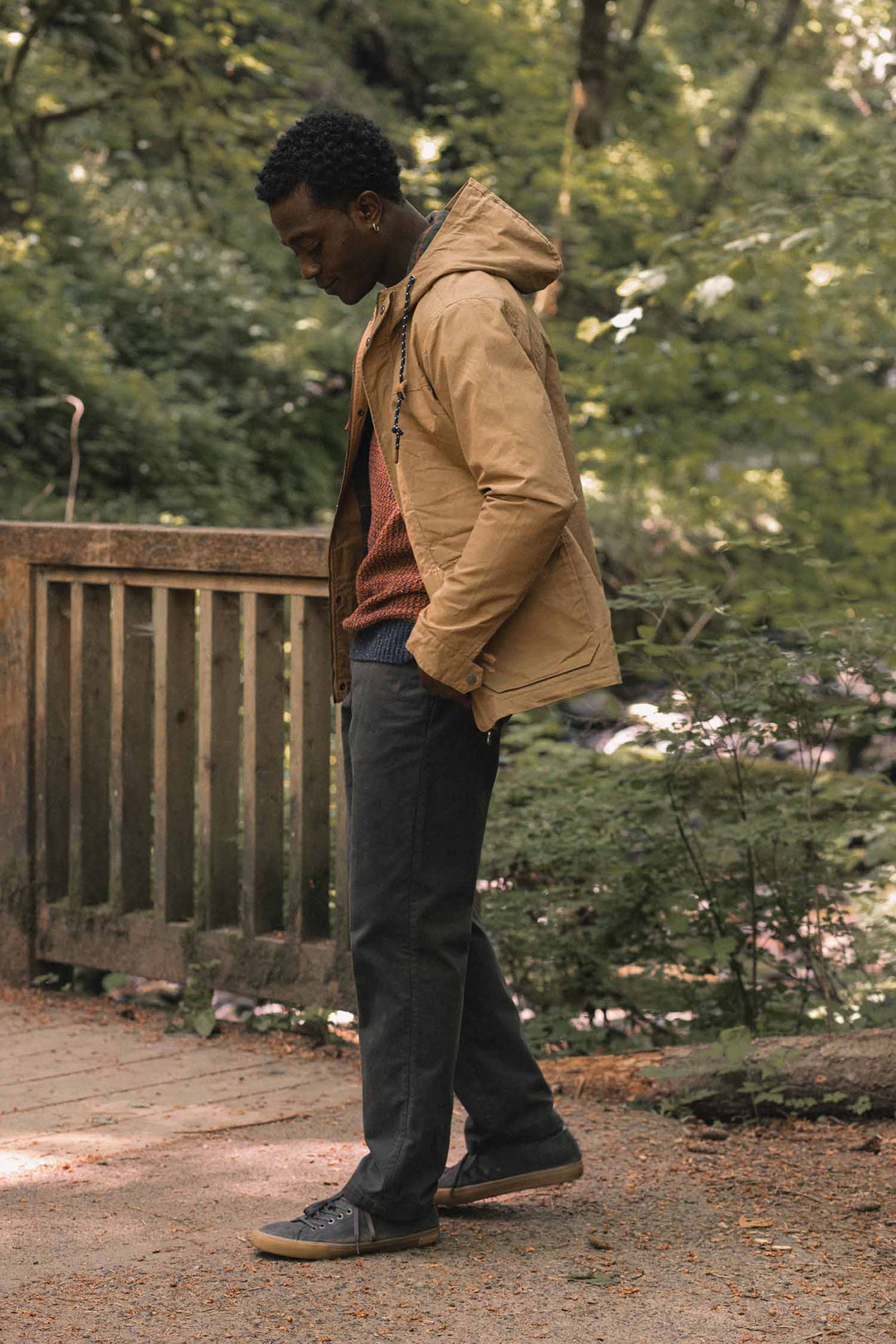 A man wearing a Bridge & Burn Marshall Jacket / Dark Khaki Waxed Canvas over an orange sweater walks across a wooden bridge.