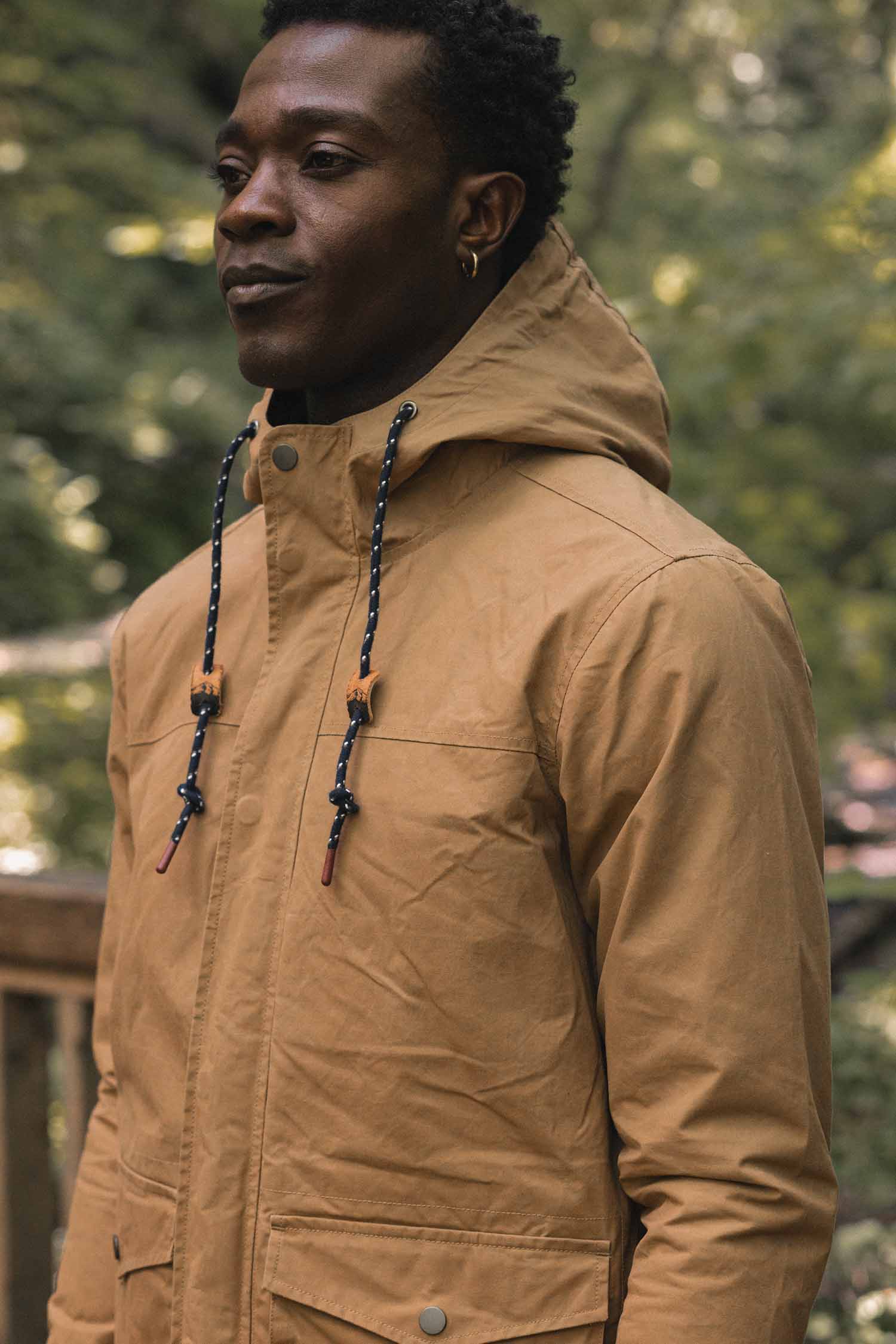 A man wearing a Bridge & Burn Marshall Jacket / Dark Khaki Waxed Canvas, featuring a hood, front zipper, snap-button closures, and patterned drawstrings.
