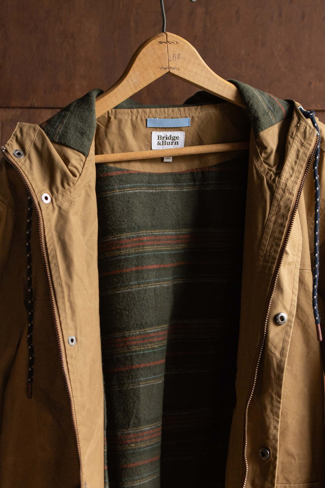 Bridge & Burn Marshall Jacket / Dark Khaki Waxed Canvas, a tan water-resistant hooded jacket with a dark green striped inner lining, hanging on a wooden hanger.