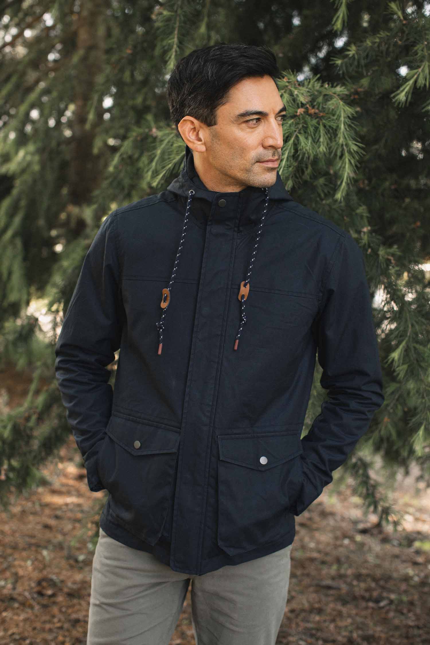 A man wearing a Bridge & Burn Marshall Jacket in navy waxed canvas, featuring a hood, two front pockets, and patterned drawstrings.