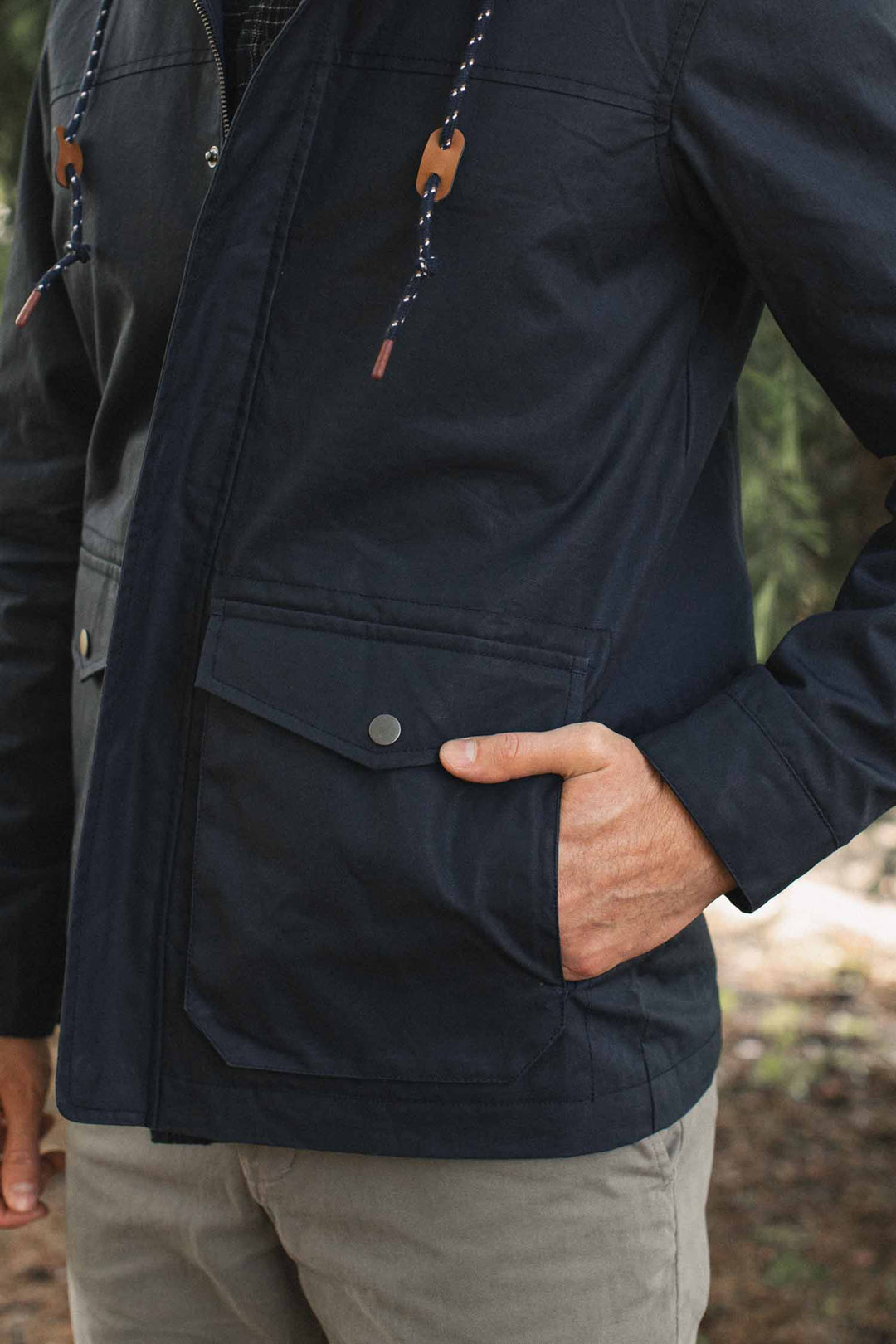 Close-up of a navy Marshall Jacket / Navy Waxed Canvas with a hood, drawstring, and snap-button flap pocket, with a hand resting in the pocket.
