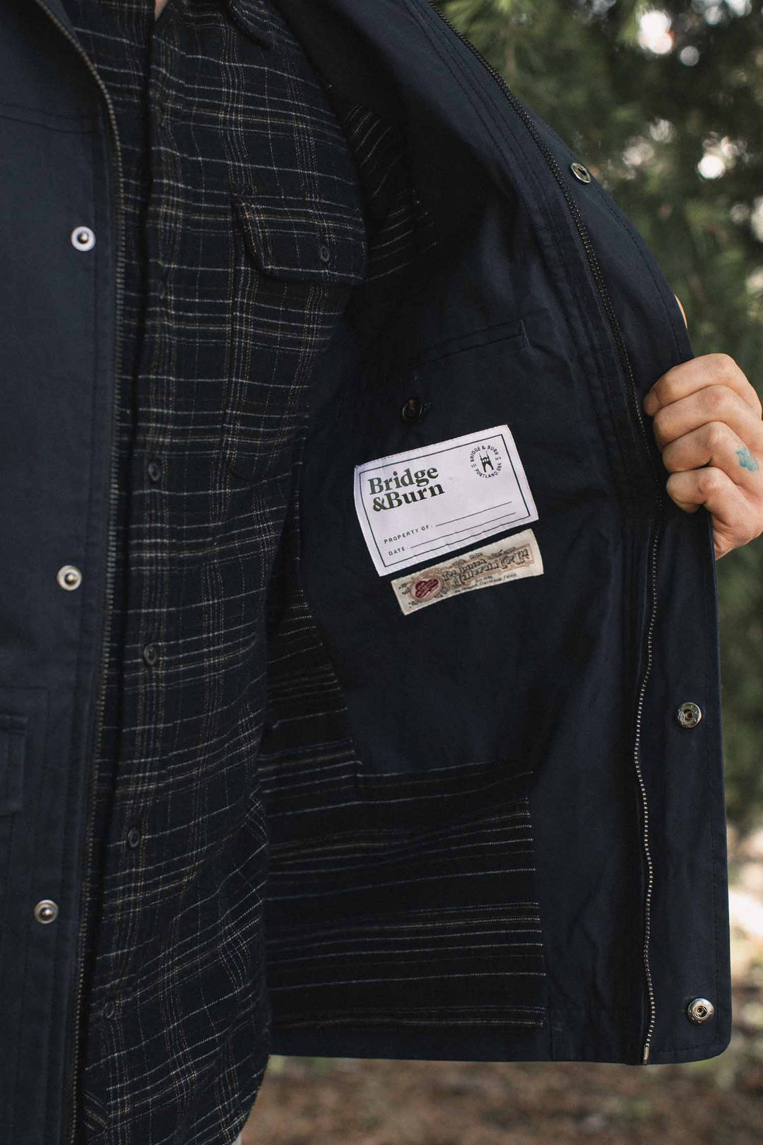 Close-up of a navy Bridge & Burn Marshall Jacket / Navy Waxed Canvas being held open to reveal the interior labels and a plaid shirt underneath.