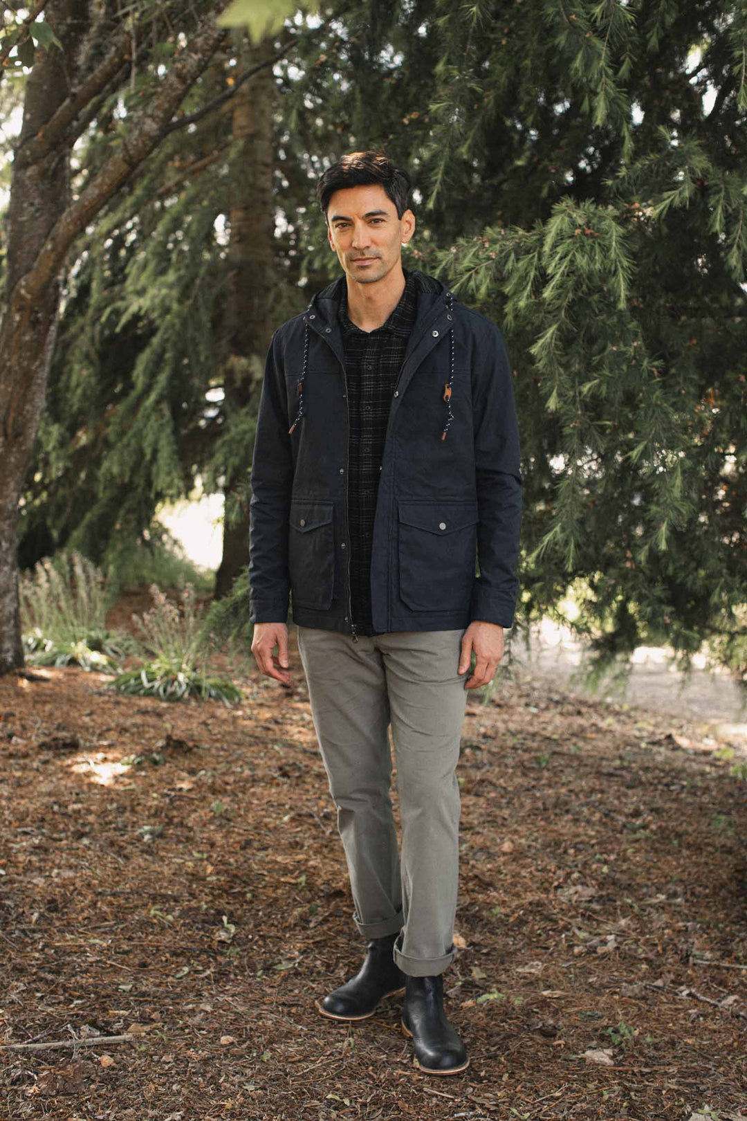 A man stands outdoors wearing a Bridge & Burn Marshall Jacket / Navy Waxed Canvas over a plaid shirt, grey trousers, and black boots.