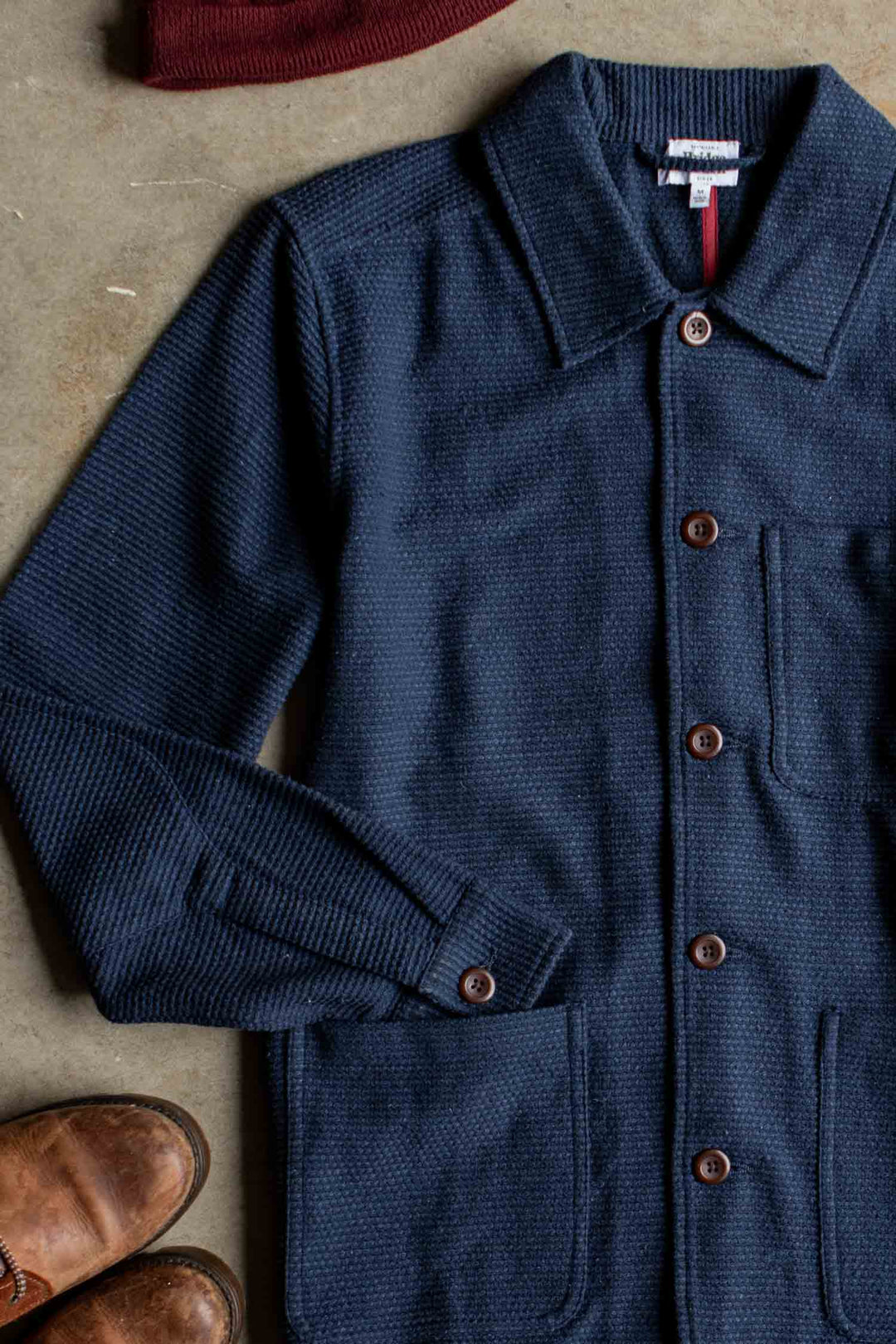 Bridge & Burn’s Men’s Studio Chore Coat in Indigo is laid flat on a beige surface, featuring distinctive brown buttons, alongside a pair of brown leather shoes and the edge of a maroon knit hat visible in the corners.