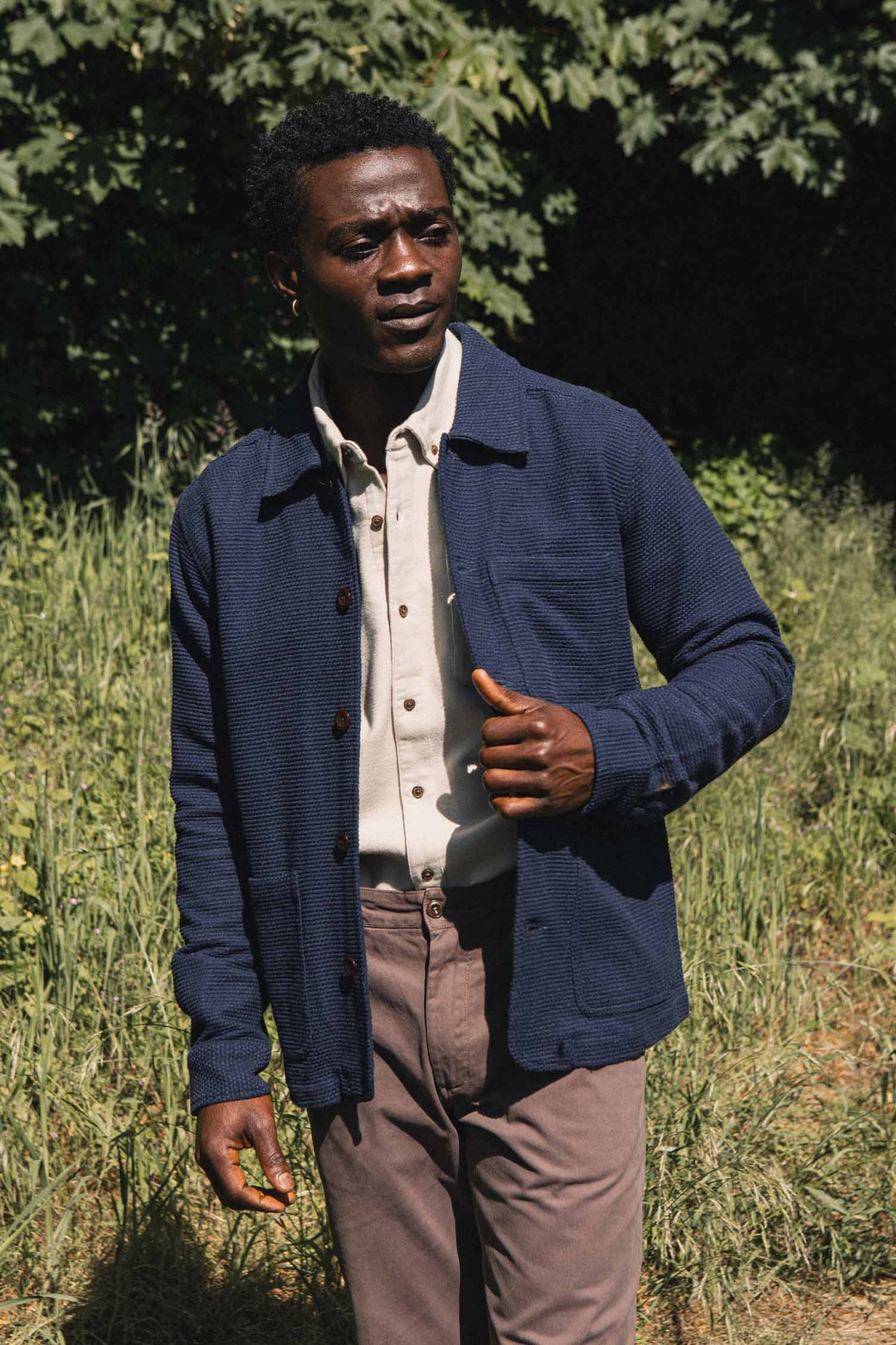 A man stands outdoors in sunlight, wearing the Bridge & Burn Men’s Studio Chore Coat in Indigo over a beige button-up shirt and mauve pants. He is in a grassy area with trees in the background, looking slightly to the side.