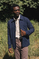 A man stands outdoors in sunlight, wearing the Bridge & Burn Men’s Studio Chore Coat in Indigo over a beige button-up shirt and mauve pants. He is in a grassy area with trees in the background, looking slightly to the side.