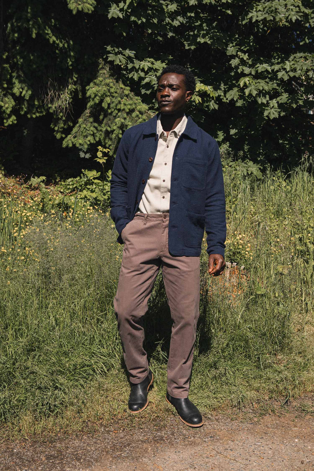 A man stands outdoors on a sunny day, wearing the Bridge & Burn Men’s Studio Chore Coat in Indigo over a cream shirt, paired with brown pants and black shoes, with greenery and trees in the background.