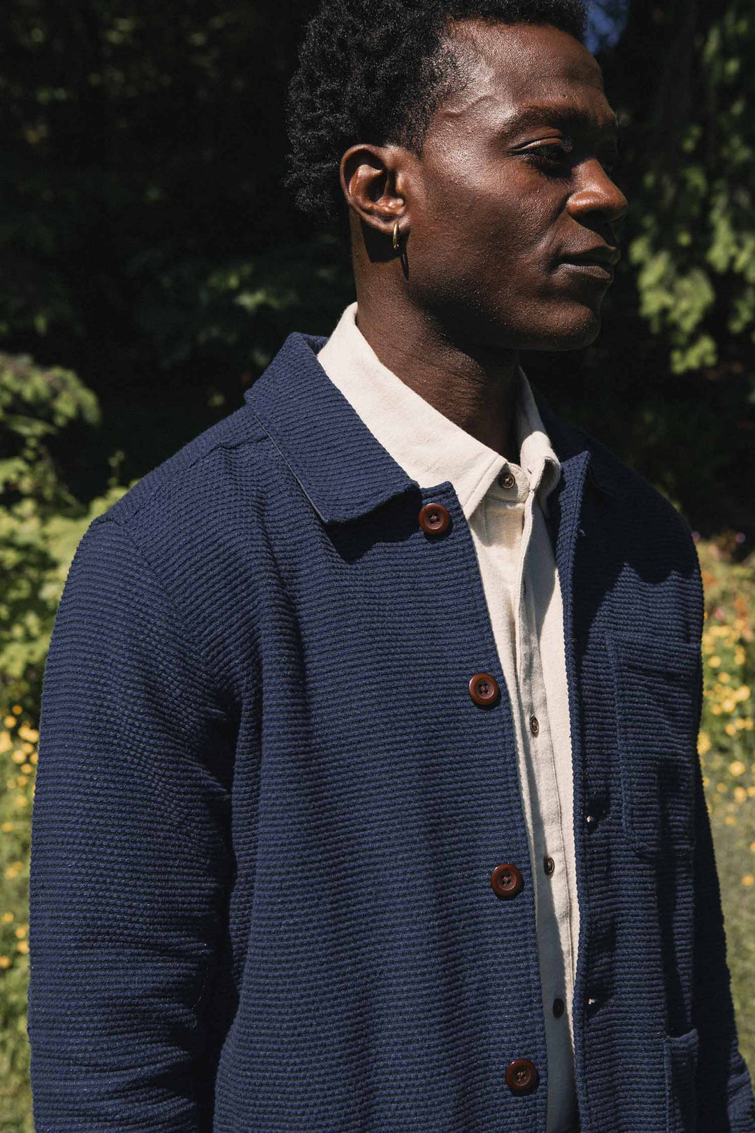 A man wearing the Bridge & Burn Men’s Studio Chore Coat in indigo over a white shirt stands outdoors in sunlight, surrounded by greenery and yellow flowers.