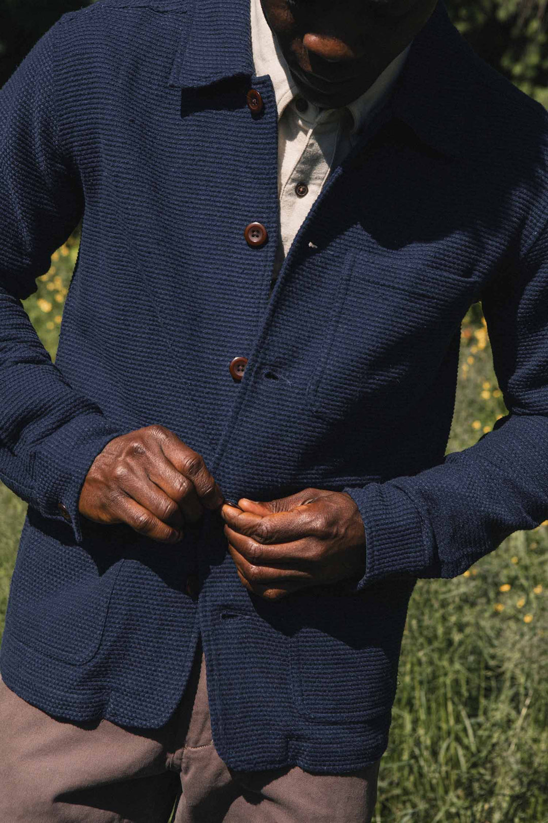 A person standing outdoors in sunlight buttons up the Bridge & Burn Men’s Studio Chore Coat in Indigo—a navy blue textured organic cotton workwear jacket—over a light-colored shirt, with green grass and yellow flowers blurred in the background.