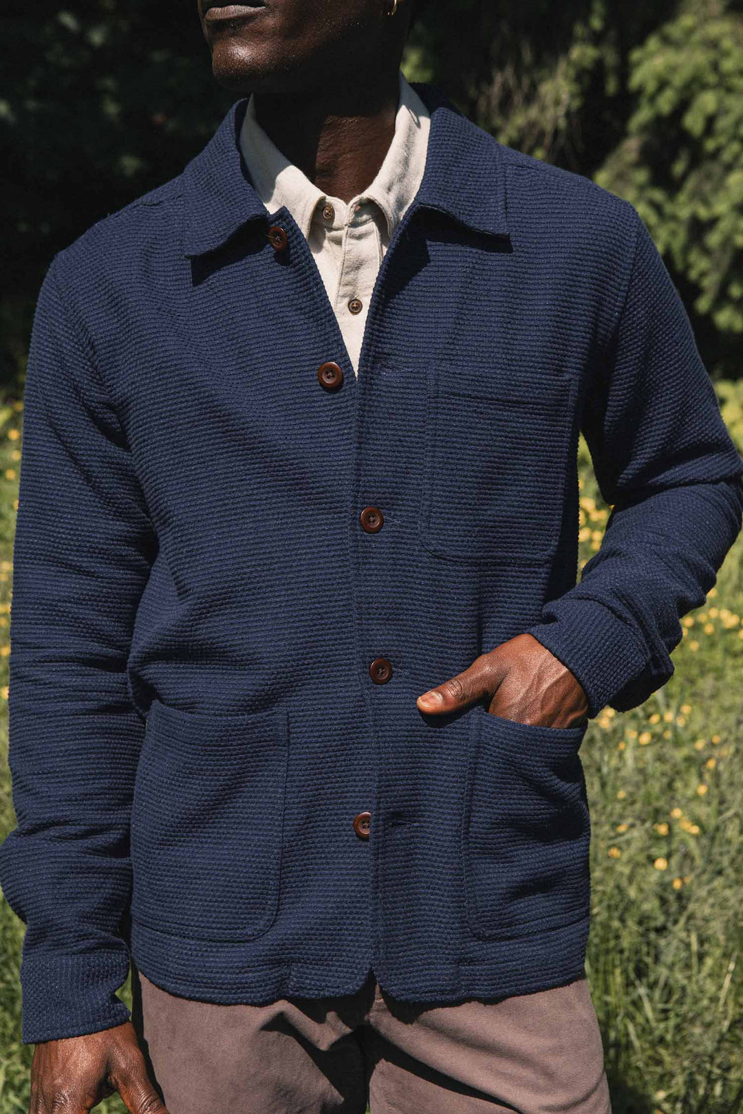 Wearing the Bridge & Burn Men’s Studio Chore Coat in Indigo over a light shirt, a person stands outdoors with one hand in their pocket, surrounded by greenery and yellow flowers.