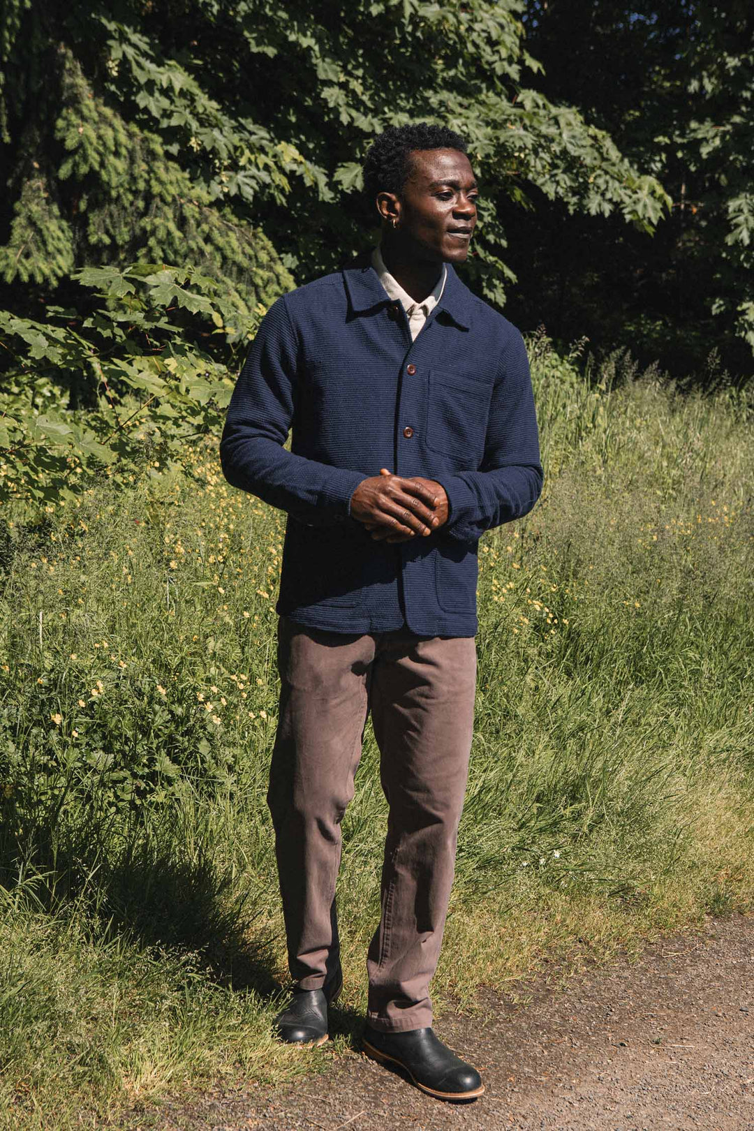 A man stands on a sunlit path by tall grass and leafy trees, wearing the Bridge & Burn Men’s Studio Chore Coat in Indigo, light brown pants, and black shoes, with his hands clasped and looking to the side.