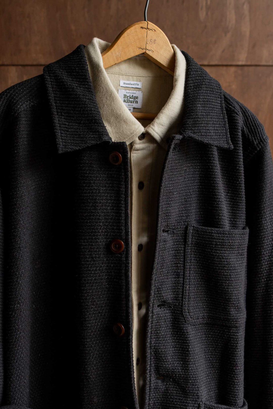 A Bridge & Burn Men’s Studio Chore Coat in washed black is layered over a beige collared shirt, both displayed on a wooden hanger against a brown wooden background.