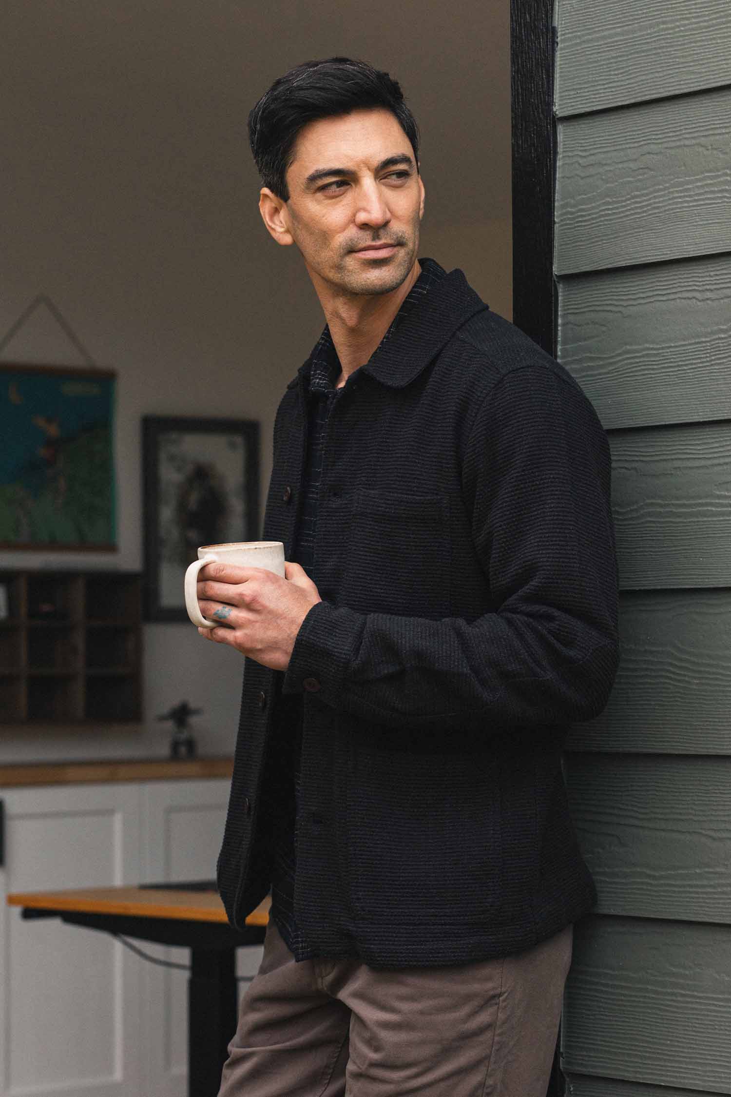 A man with dark hair stands outside near a building, holding a white mug and looking to the side. He wears the Bridge & Burn Men’s Studio Chore Coat in Washed Black over a dark shirt and tan pants. In the background, part of a room with artwork on the wall is visible.