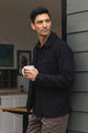 A man with dark hair stands outside near a building, holding a white mug and looking to the side. He wears the Bridge & Burn Men’s Studio Chore Coat in Washed Black over a dark shirt and tan pants. In the background, part of a room with artwork on the wall is visible.