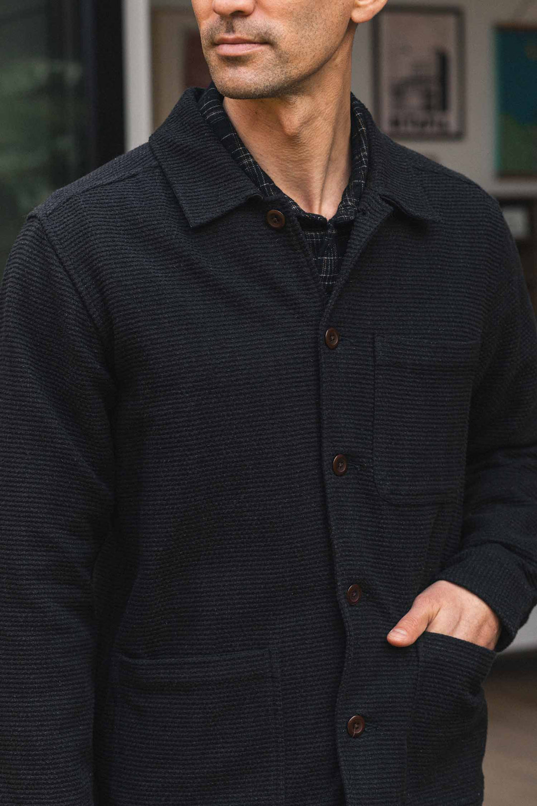 A man wearing the Bridge & Burn Men’s Studio Chore Coat in Washed Black, featuring brown buttons, stands with one hand in his pocket. His face is partially visible, and blurred framed artwork appears in the background.