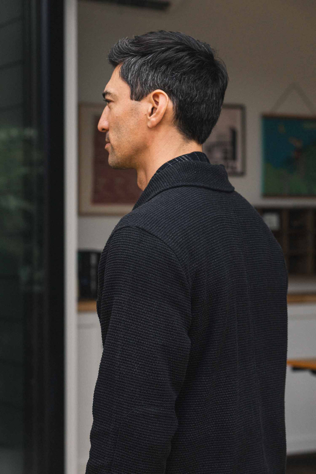 A man with short dark hair and a black textured sweater stands indoors, facing left. The blurred wall art and light-toned shelves in the background evoke the relaxed ambiance of a studio or loft—his look subtly complemented by the Men’s Studio Chore Coat in Washed Black from Bridge & Burn, a modern workwear jacket thoughtfully crafted from organic cotton.