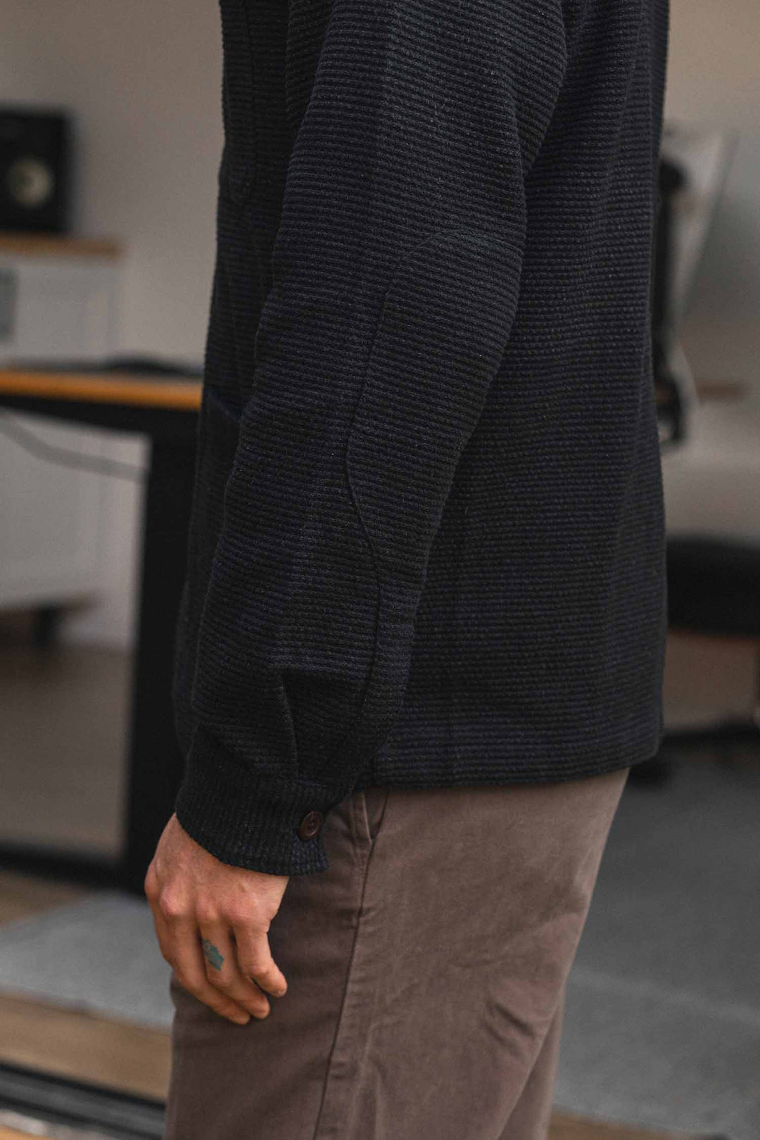 A person stands indoors, wearing the Bridge & Burn Men’s Studio Chore Coat in Washed Black paired with light brown pants and their arm relaxed by their side. The background shows a desk and office chair, complementing the subtle workwear jacket aesthetic of the coat.