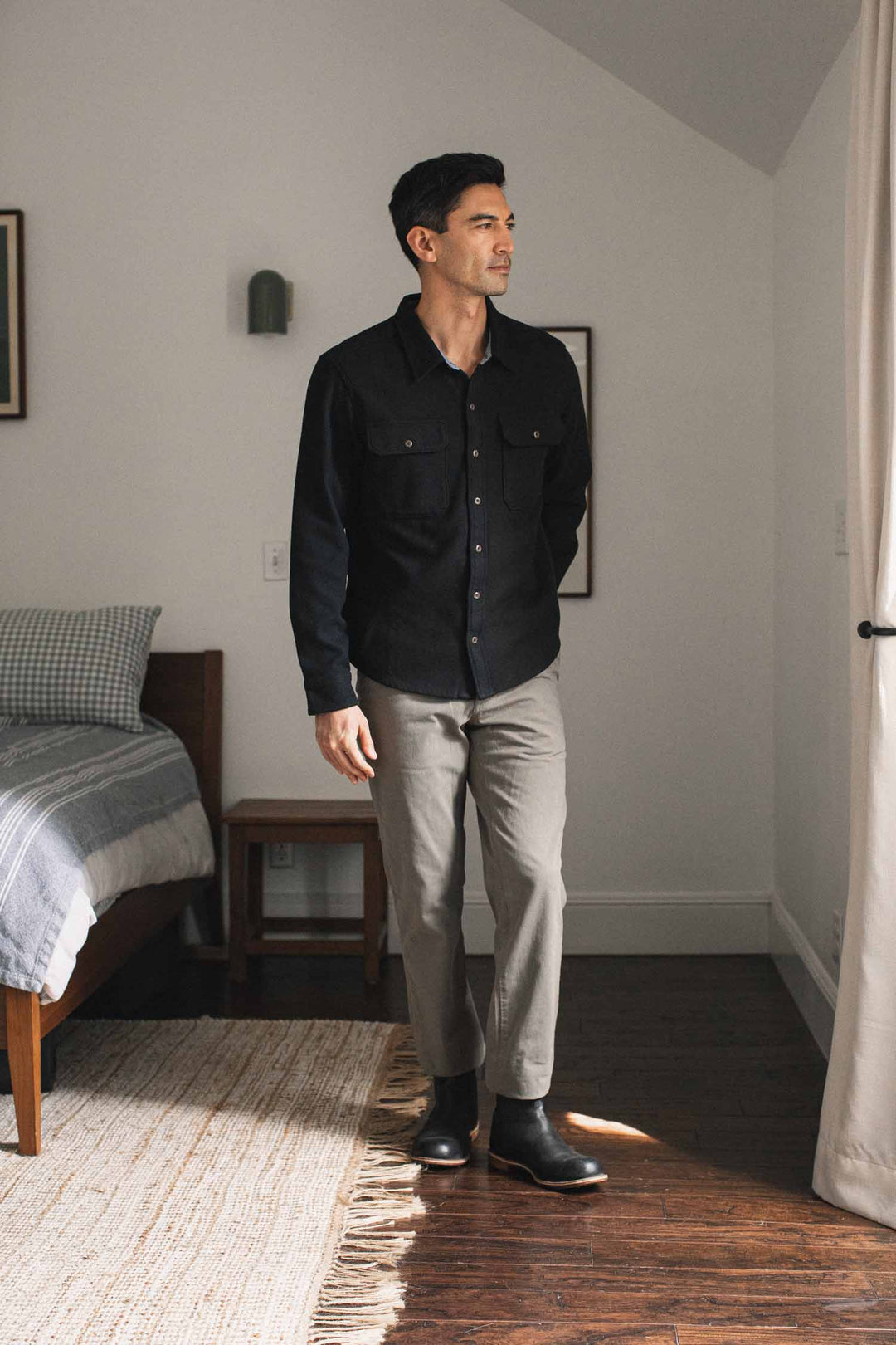 A man in the Pointer Overshirt in Ink Stone from Bridge & Burn, paired with gray pants and black boots, stands in a bright bedroom furnished with a bed, rug, and curtains. His outfit reflects a commitment to sustainable clothing, combining style with eco-conscious choices.
