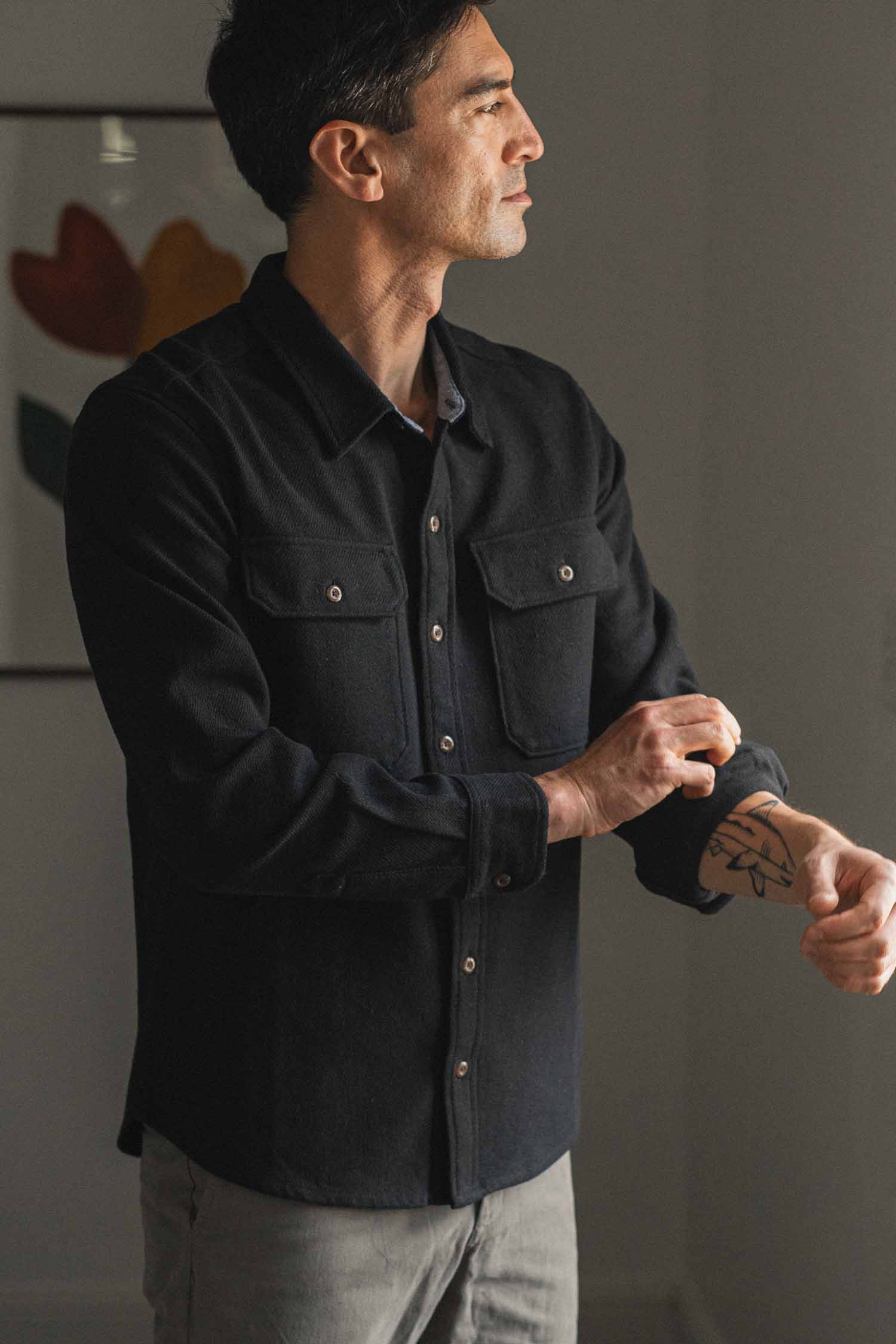 A man with dark hair, wearing the Bridge & Burn Pointer Overshirt in Ink Stone and gray pants, stands indoors rolling up his sleeve to reveal a tattoo on his forearm. A framed abstract artwork is visible in the background.