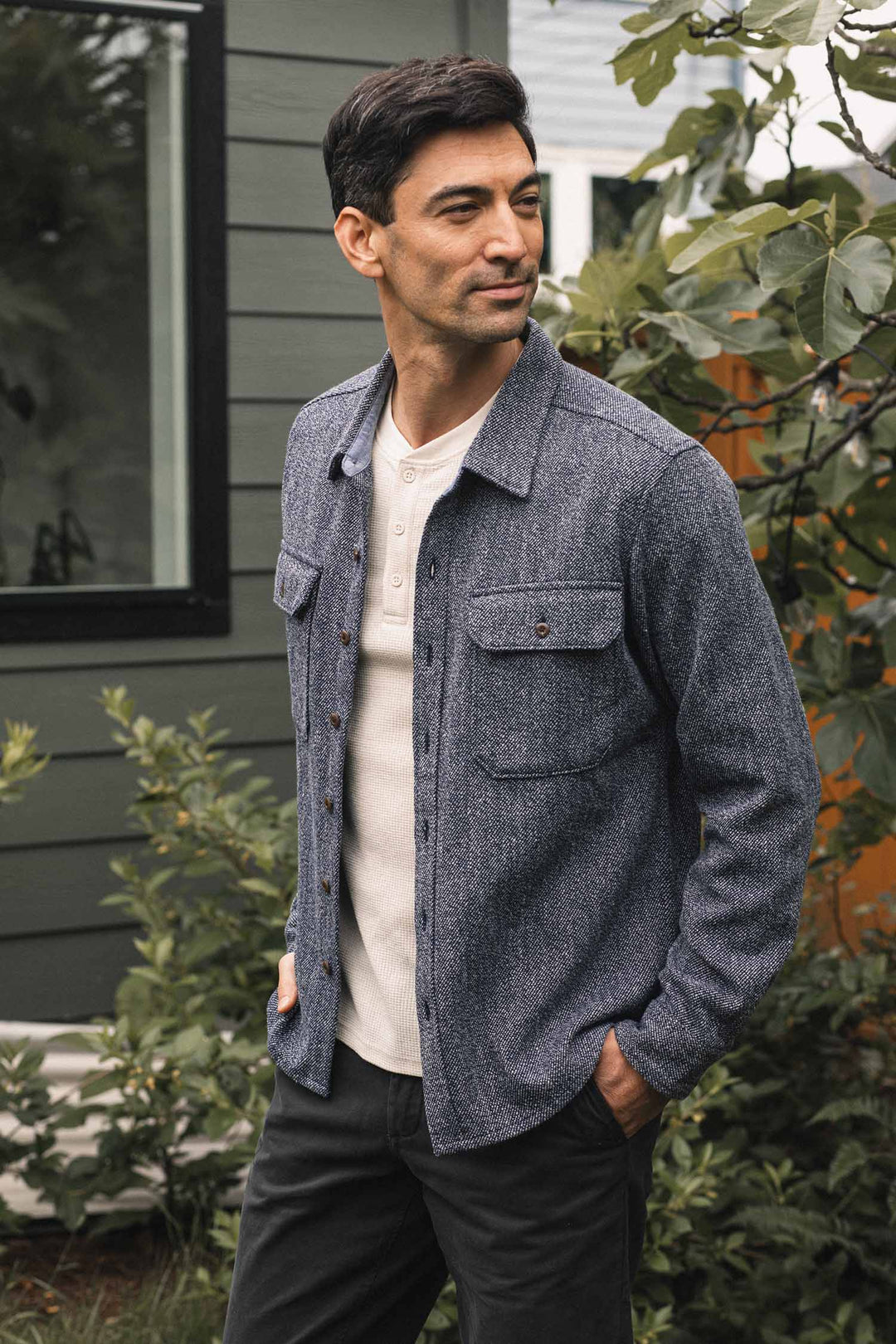 A man stands outdoors near a green house, wearing the Bridge & Burn Pointer Overshirt in Ocean Marl—a blue twill overshirt made from recycled fibers—layered over a white henley and dark pants, with his hands in his pockets, looking to the side.