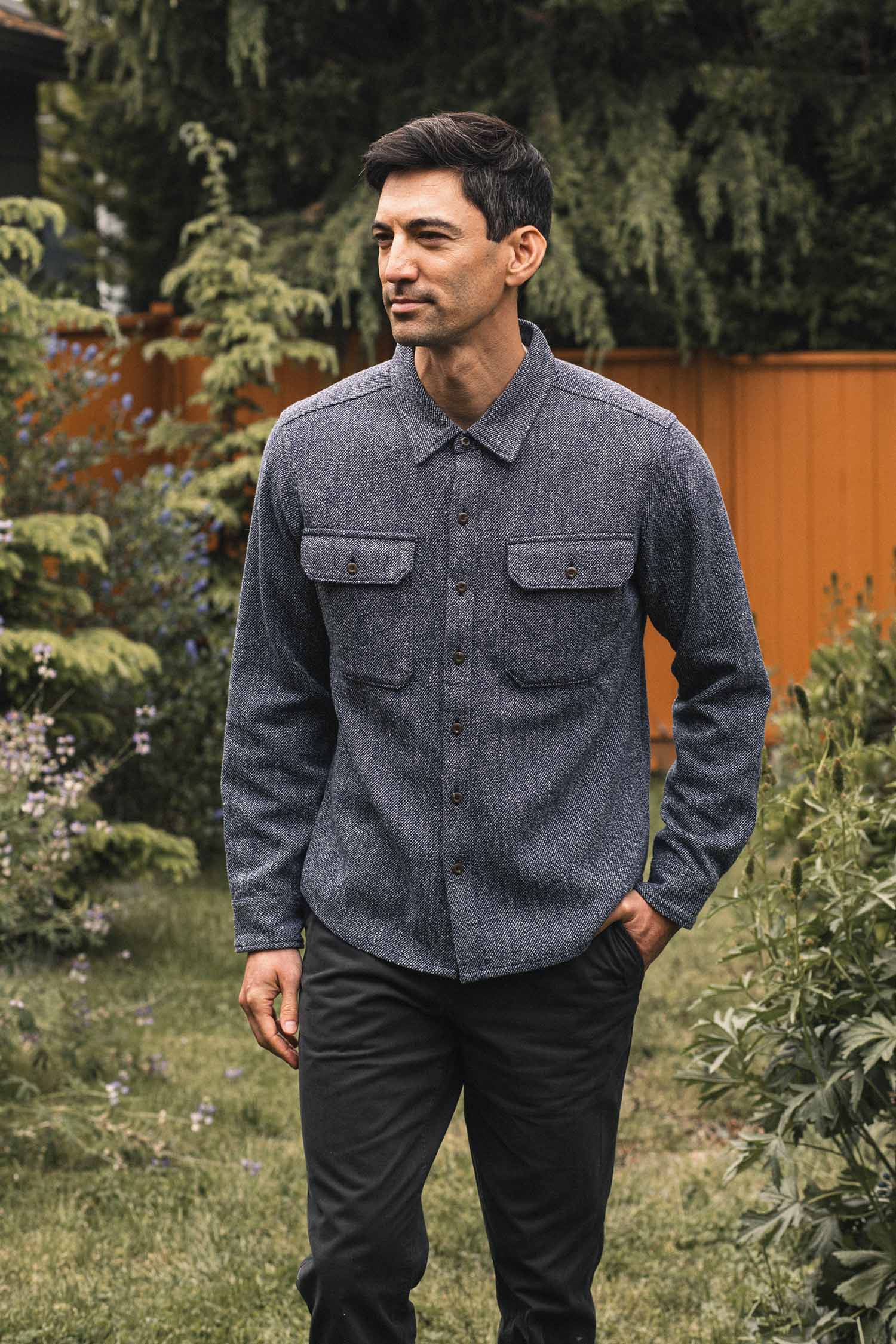 A man wearing the Bridge & Burn Pointer Overshirt in Ocean Marl and black pants stands outside in a garden with green plants and trees, with an orange fence in the background.