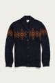 The Sprague Cardigan in Highland Dusk by Bridge & Burn is a navy blue shawl collar cardigan crafted from soft RWS wool, featuring a button closure, ribbed trim, front pockets, and an orange geometric pattern across the chest and sleeves.