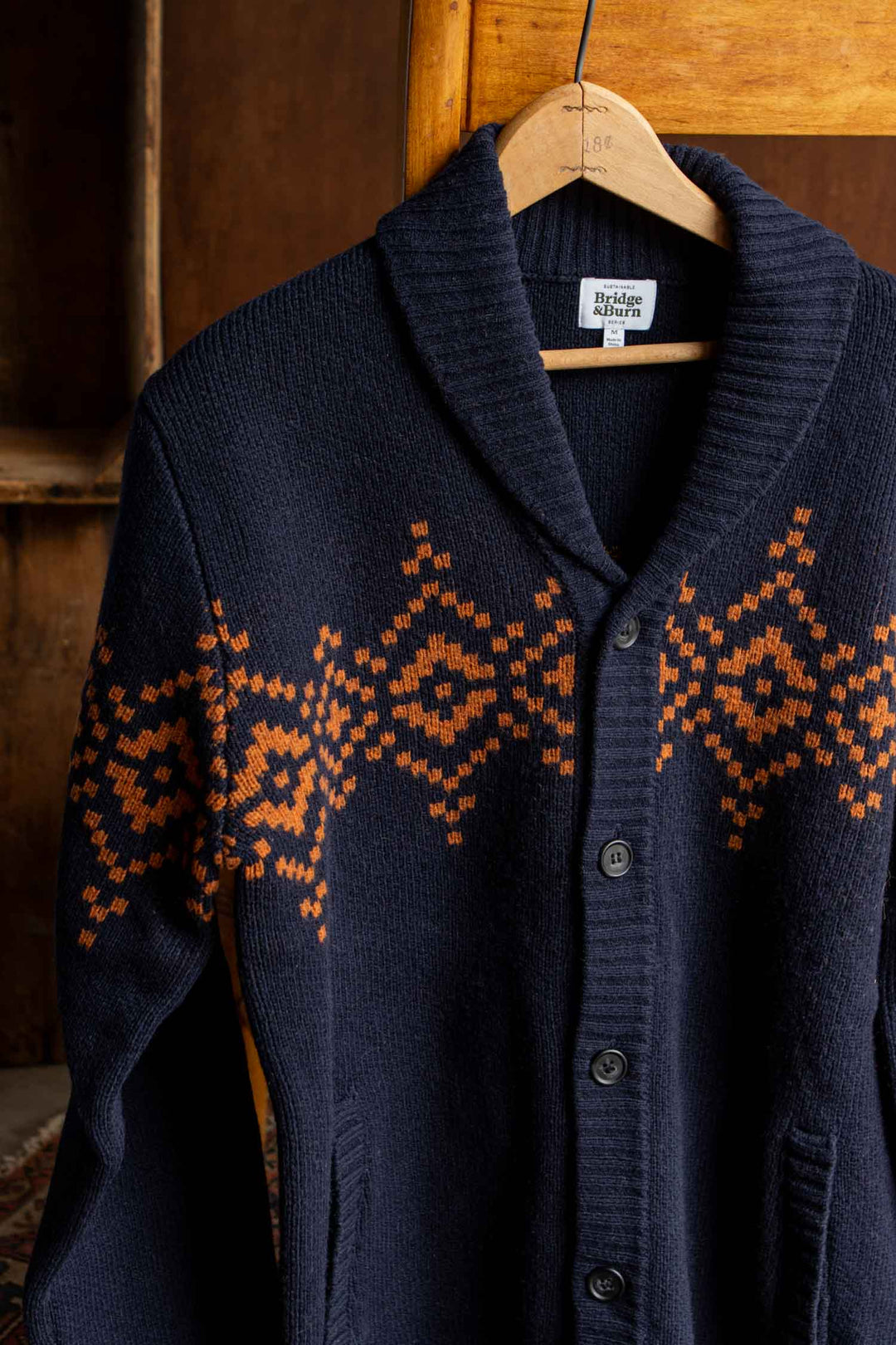 The Sprague Cardigan in Highland Dusk by Bridge & Burn is displayed on a wooden hanger against a wooden background. Crafted from soft RWS wool, this navy blue knit features orange geometric patterns across the chest and sleeves and is finished with ribbed trim for a polished look.