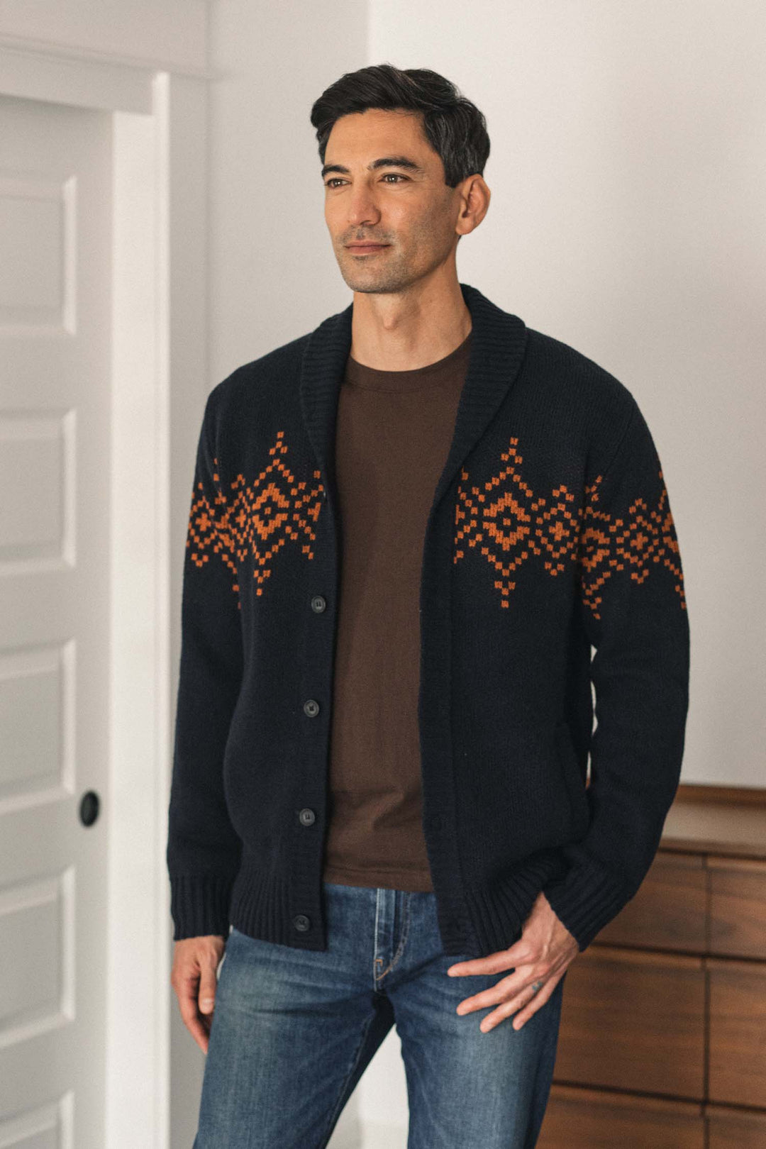 A man wearing the Bridge & Burn Sprague Cardigan in Highland Dusk—a dark RWS wool shawl collar cardigan with an orange geometric pattern and ribbed trim—pairs it with a brown T-shirt and jeans as he stands indoors near a white door and wooden cabinet.