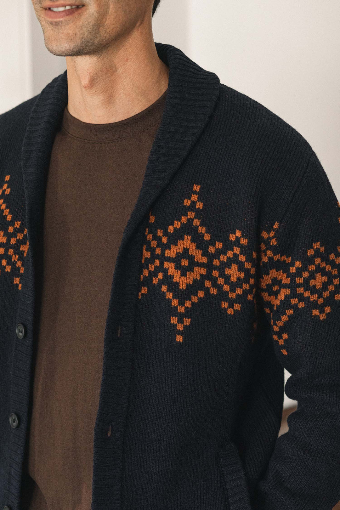 A person wearing the Bridge & Burn Sprague Cardigan in the Highland Dusk colorway, featuring orange geometric patterns and ribbed trim over a brown shirt, shown from the shoulders to the waist.
