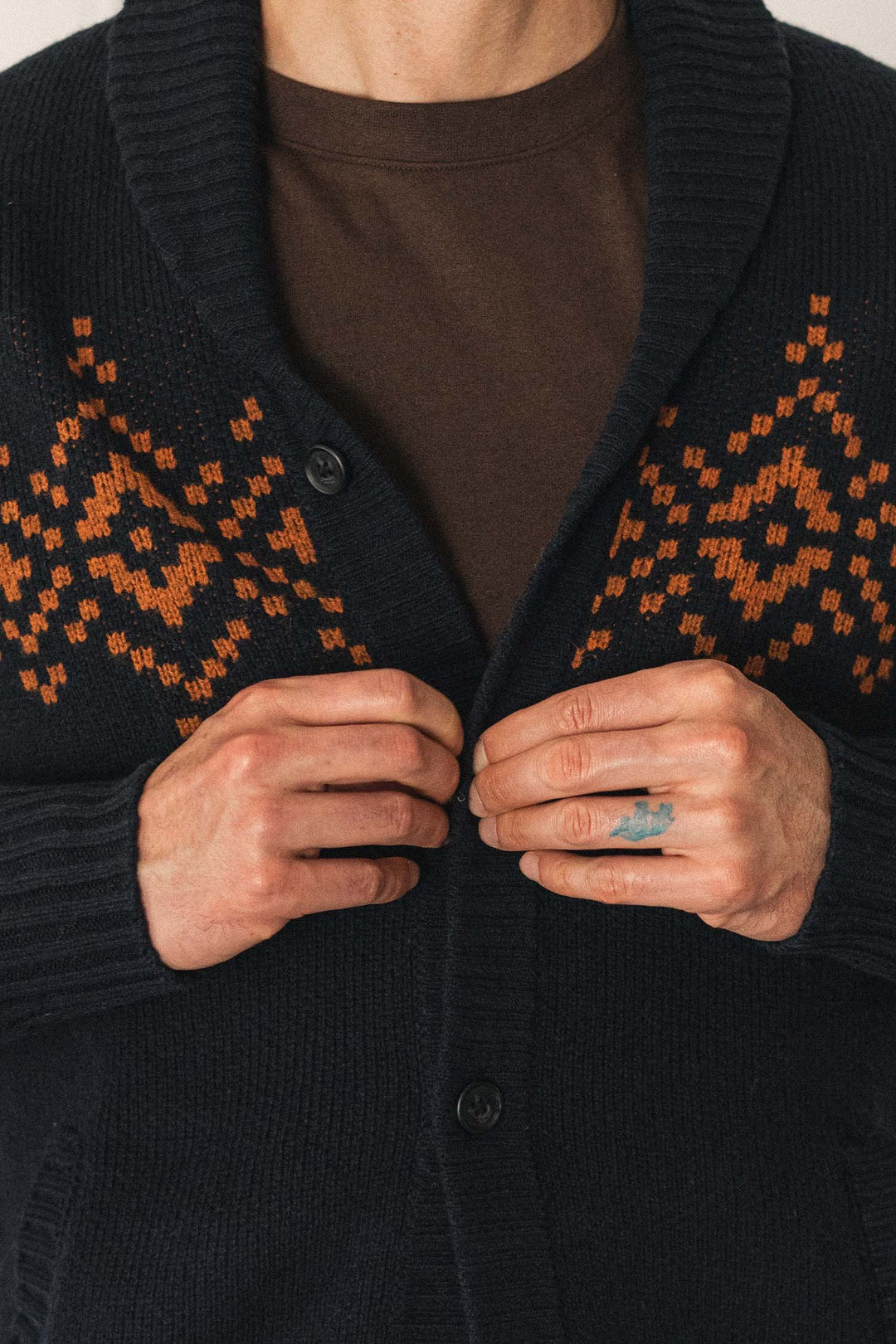 A person wearing the Bridge & Burn Sprague Cardigan in Highland Dusk—a dark shawl collar sweater featuring an orange geometric pattern—over a brown shirt, holding the ribbed trim edges with both hands.
