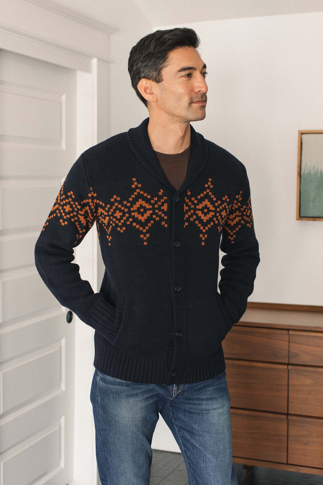 A man wearing the Bridge & Burn Sprague Cardigan in Highland Dusk, featuring a navy shawl collar and orange geometric pattern with ribbed trim, stands indoors with his hands in his pockets near a dresser and a white door.