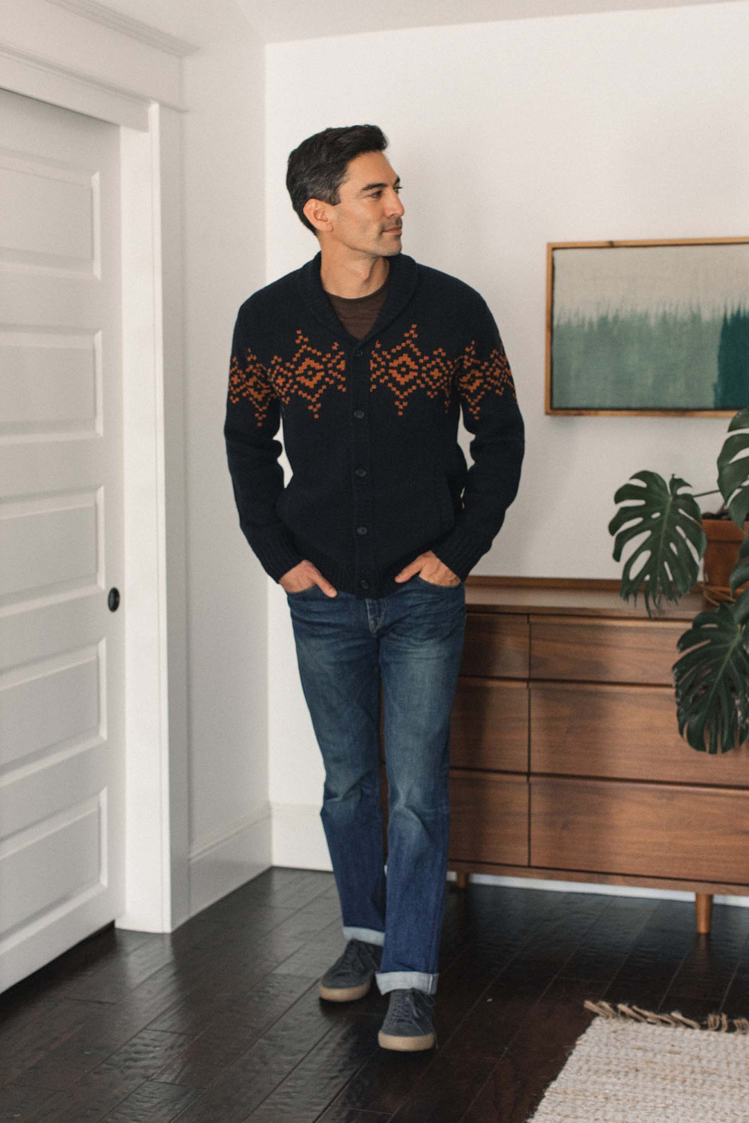 A man wearing the Bridge & Burn Sprague Cardigan in Highland Dusk, styled with blue jeans and gray shoes, stands indoors near a mid-century dresser, a plant, and abstract wall art.